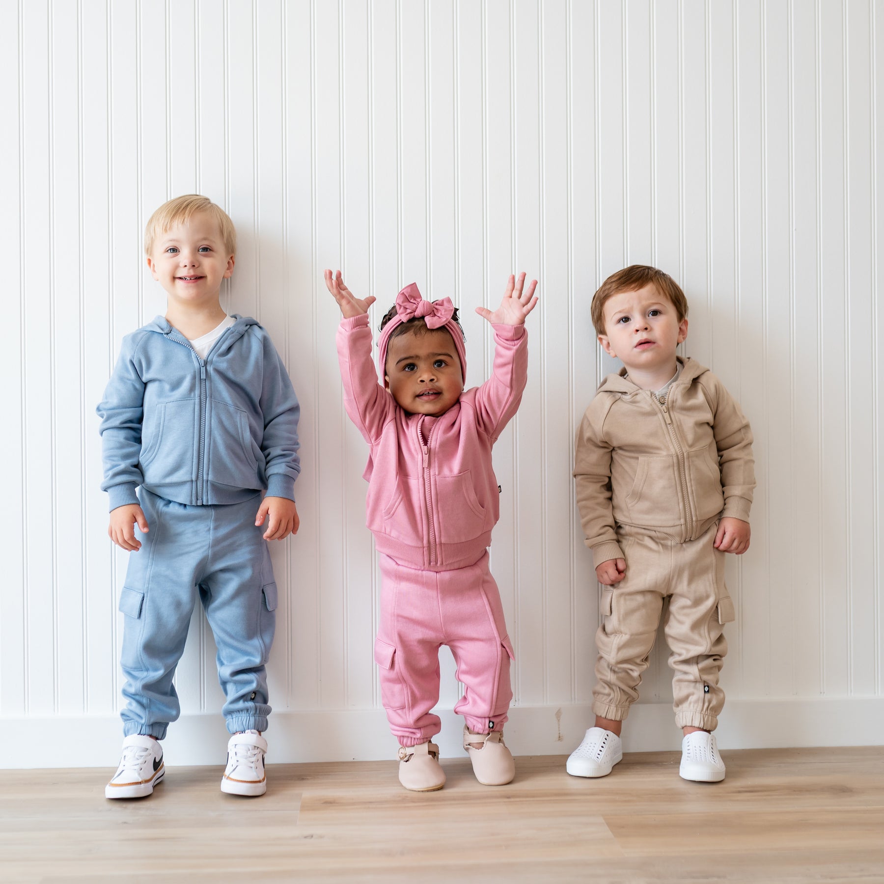 Three young kids standing in front of a white paneled wall side by side wearing the Fleece Cargo Pants in Almond, Apple Blossom and Slate with matching fleece zip hoodies
