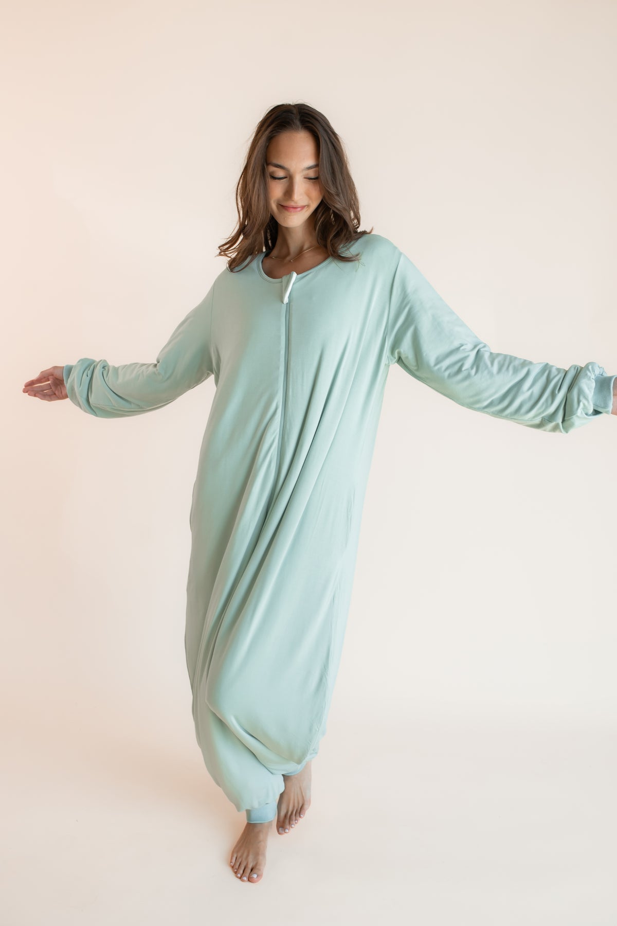 A woman wearing a light mint green full-body zip-up onesie pajama, standing barefoot against a solid pale beige background with her arms outstretched.