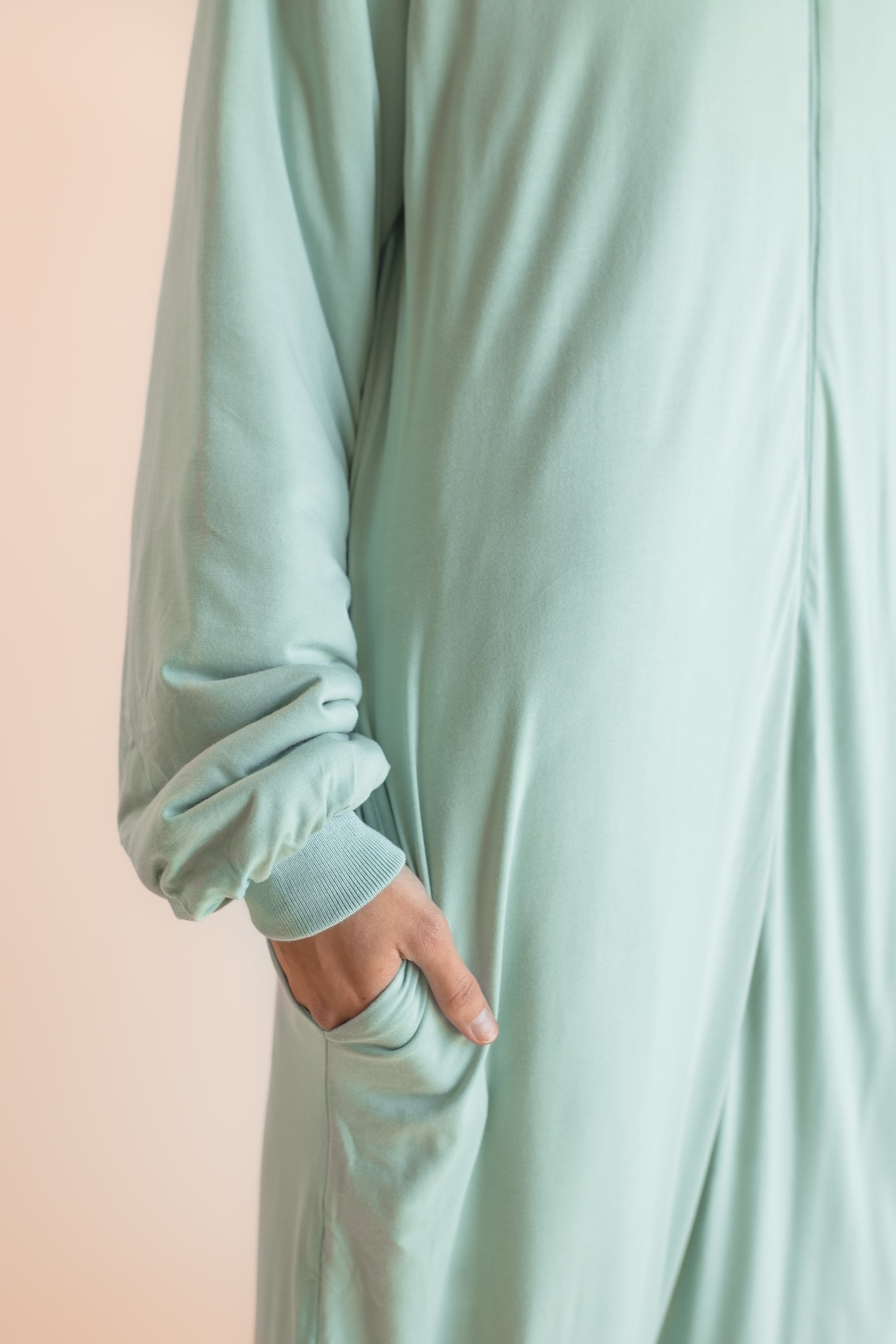 A close-up of a person wearing a long, flowing sage green garment with a gathered cuff and a side pocket, with their hand tucked inside. The background is a soft, solid pale peach-pink.