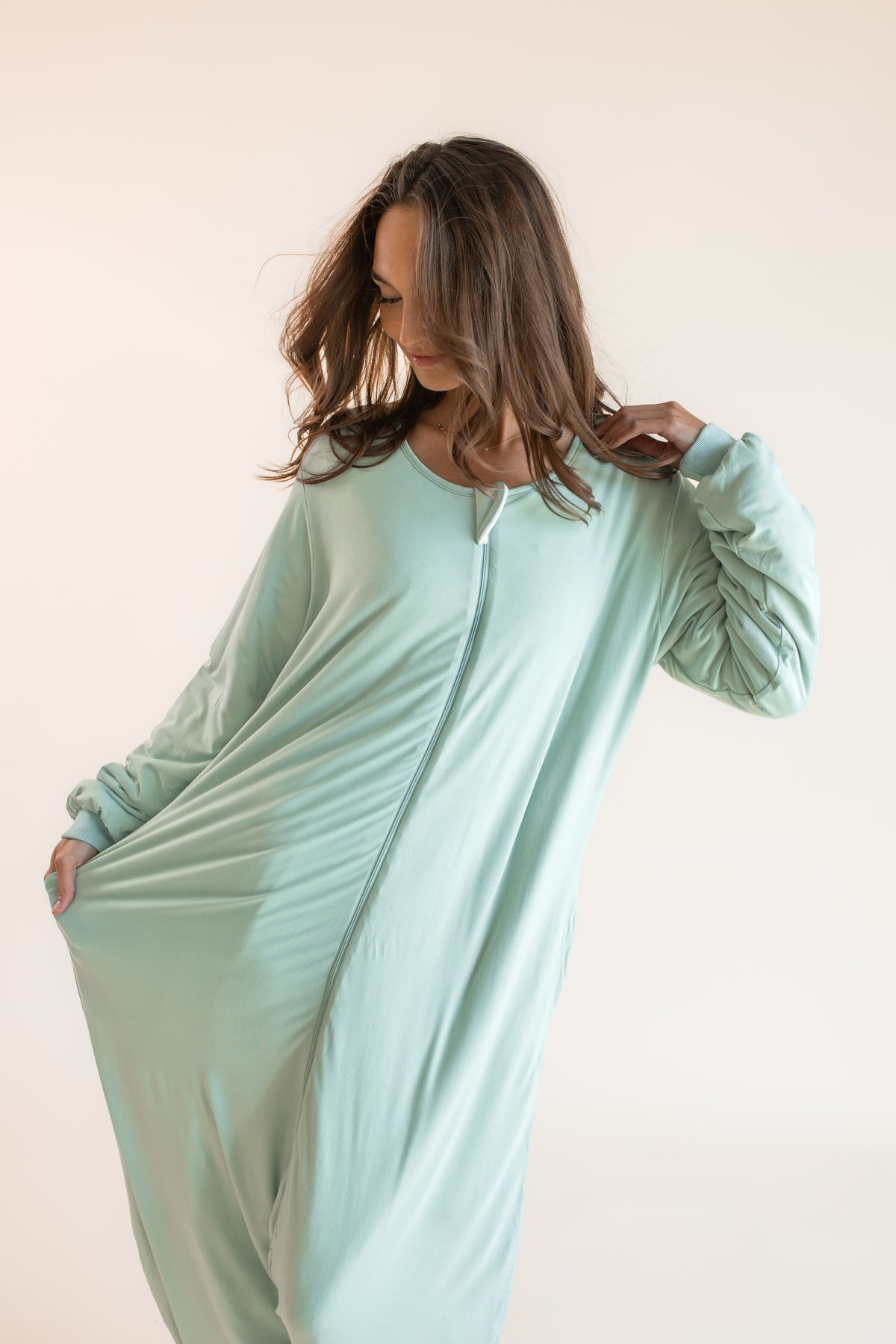 A woman wearing a long-sleeve mint green zip-up onesie pajama, posing against a solid light beige background.