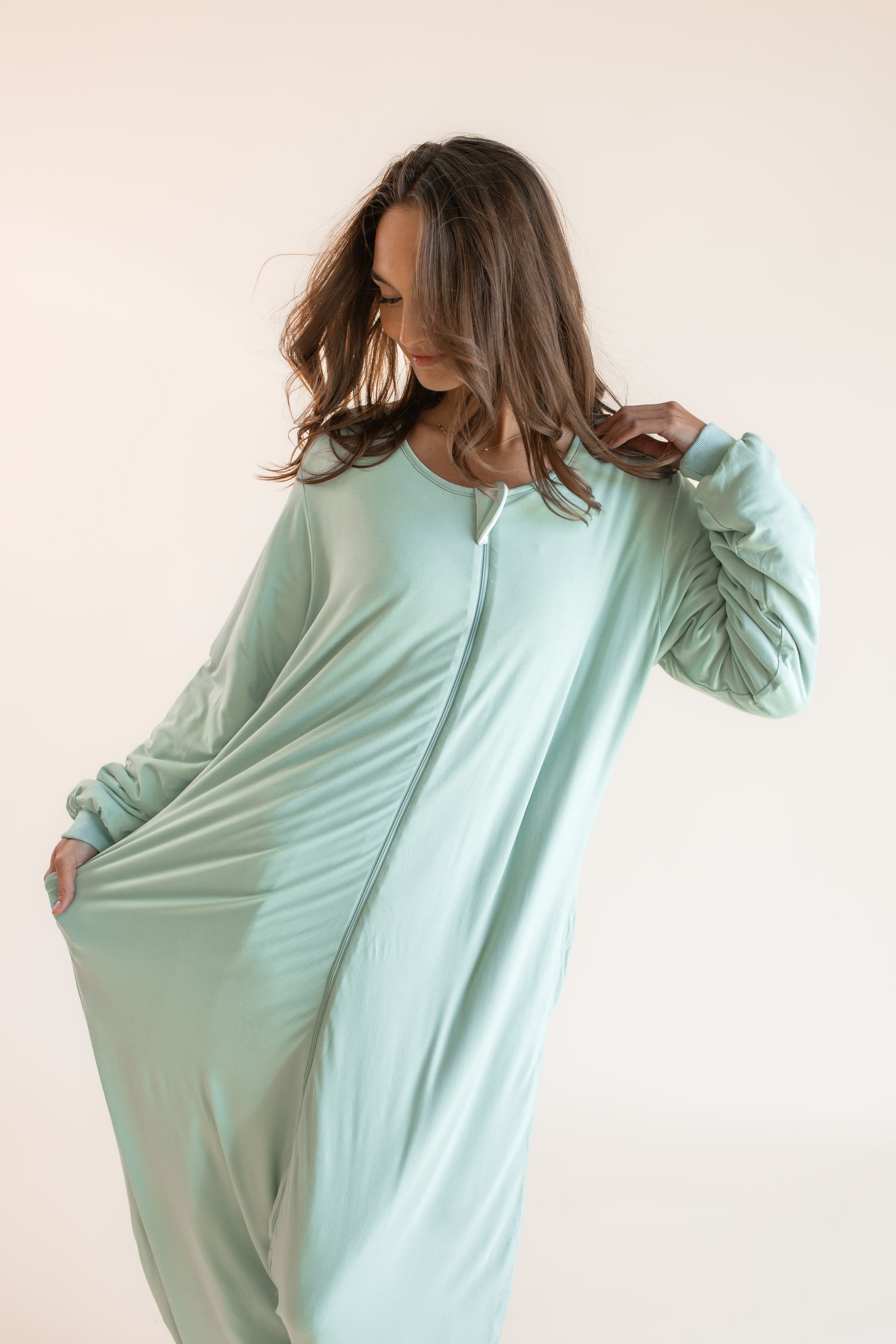A woman wearing a long-sleeve mint green zip-up onesie pajama, posing against a solid light beige background.