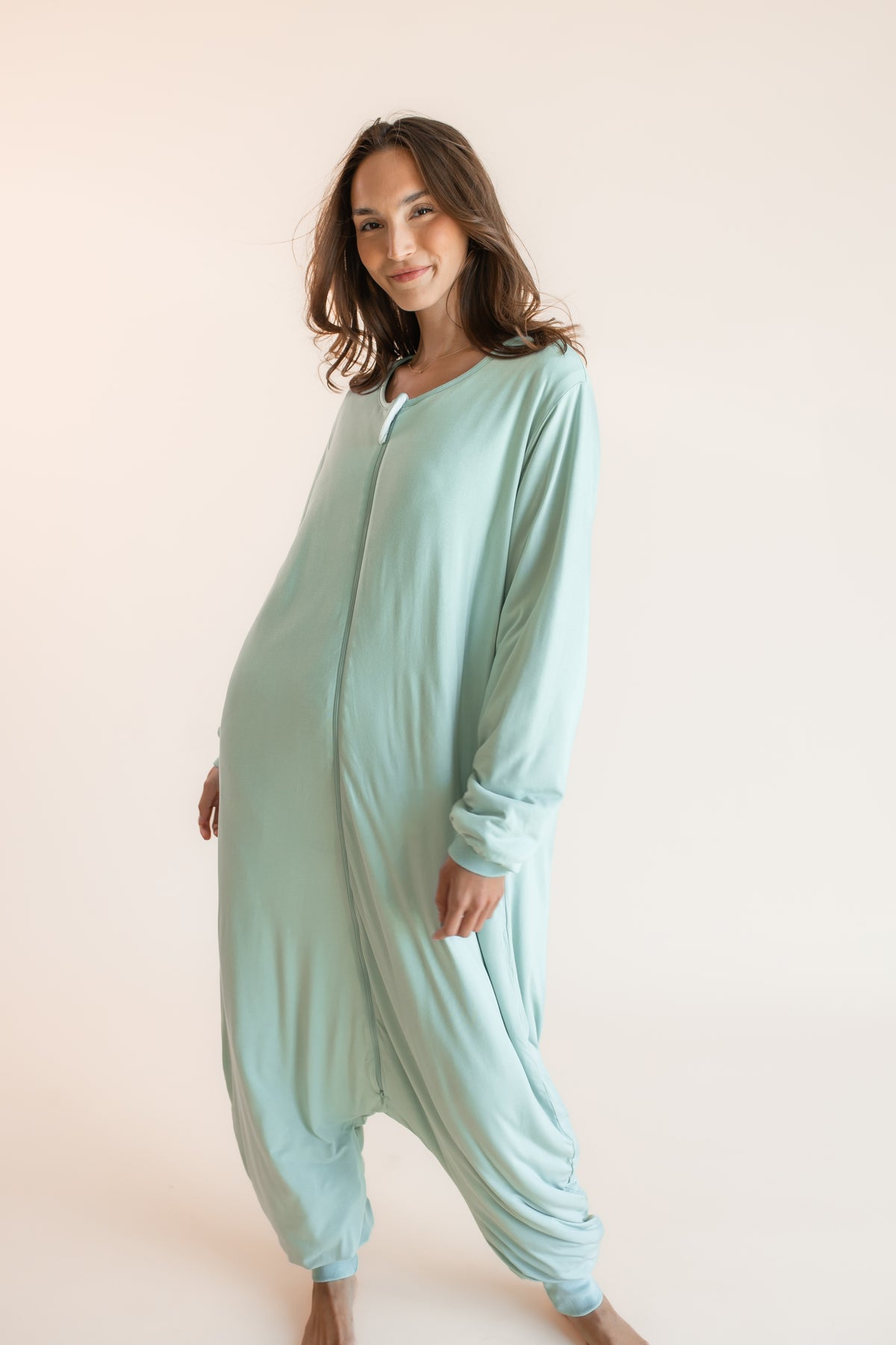 A woman modeling a light mint green, one-piece harem-style sleep sack against a solid pale peach studio background.