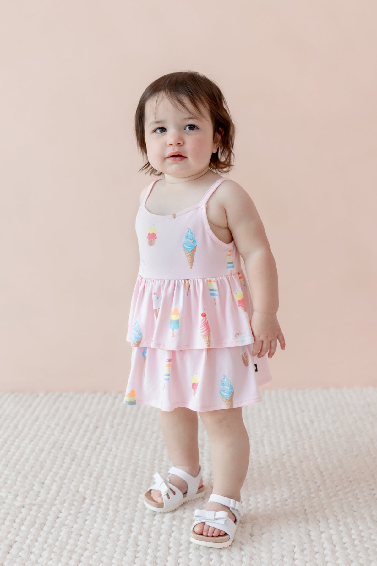 Child standing in Sakura Ice Cream print spaghetti strap ruffle romper with white sandles on beige background