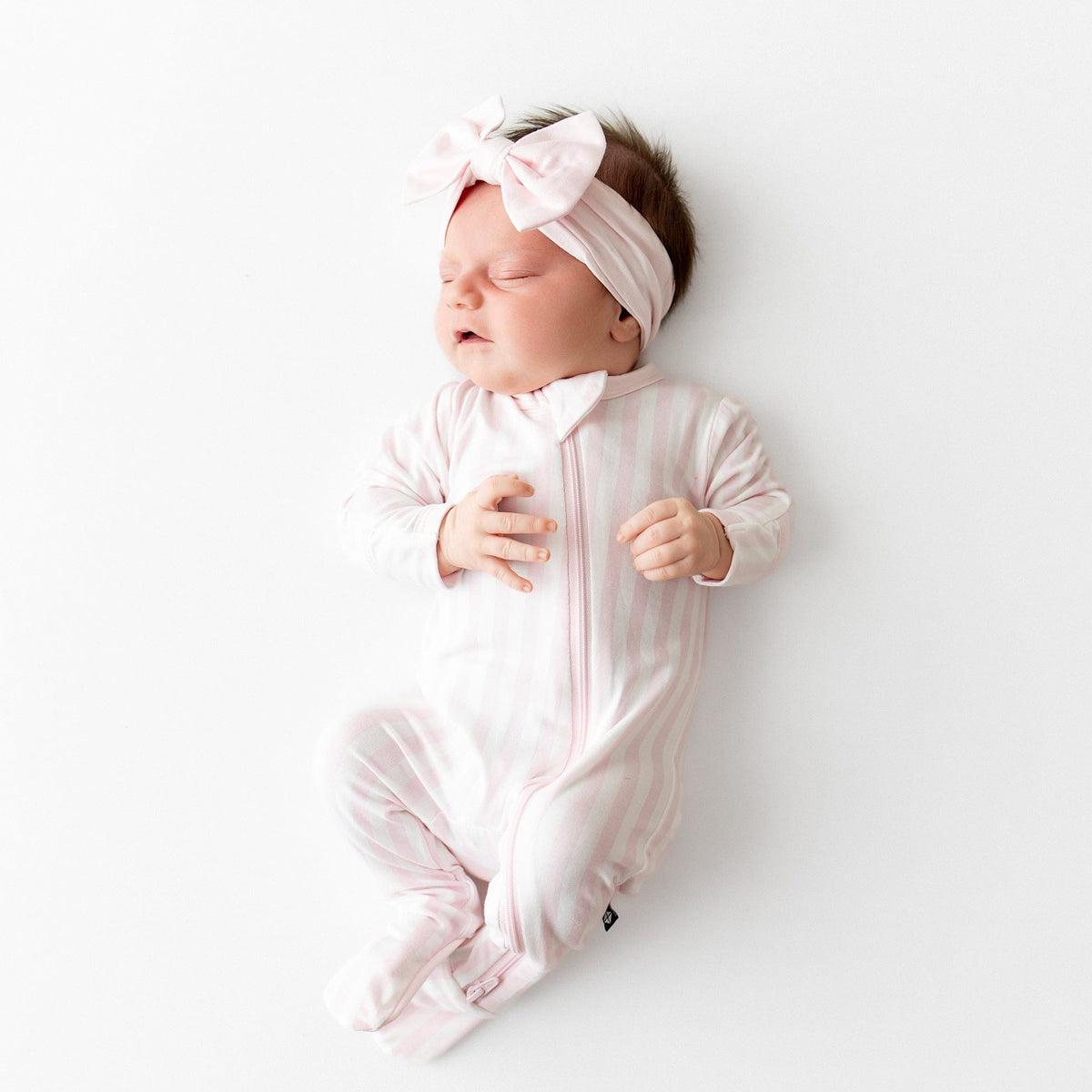 Sleeping newborn wearing the Zippered Footie in Small Sakura Stripe and Light pink bow headband 