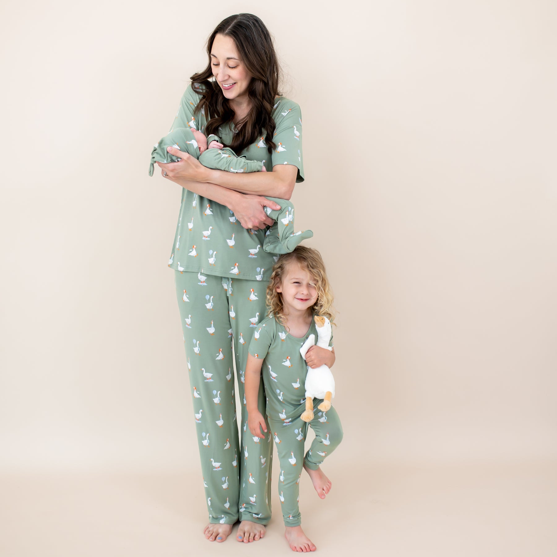 Family matching in Silly Goose. Mother wearing the Women's V-Neck and Lounge pants holding her newborn in a take me home set with Cap with her oldest child standing beside her wearing the short sleeve toddler pajamas with pant holding a toy stuffed goose