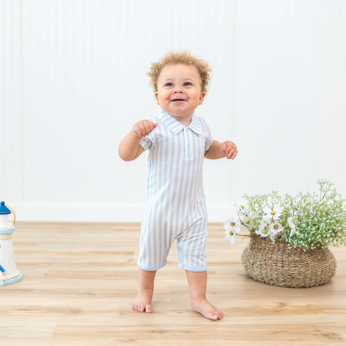 Smiling infant standing beside potted plants and flowers wearing the Polo Shortall in Small Mist Stripe