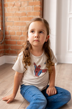 Girl sitting on floor in front of a brick wall wearing oat color Toddler Basic Tee with Spider-Man print in center over blue jeans