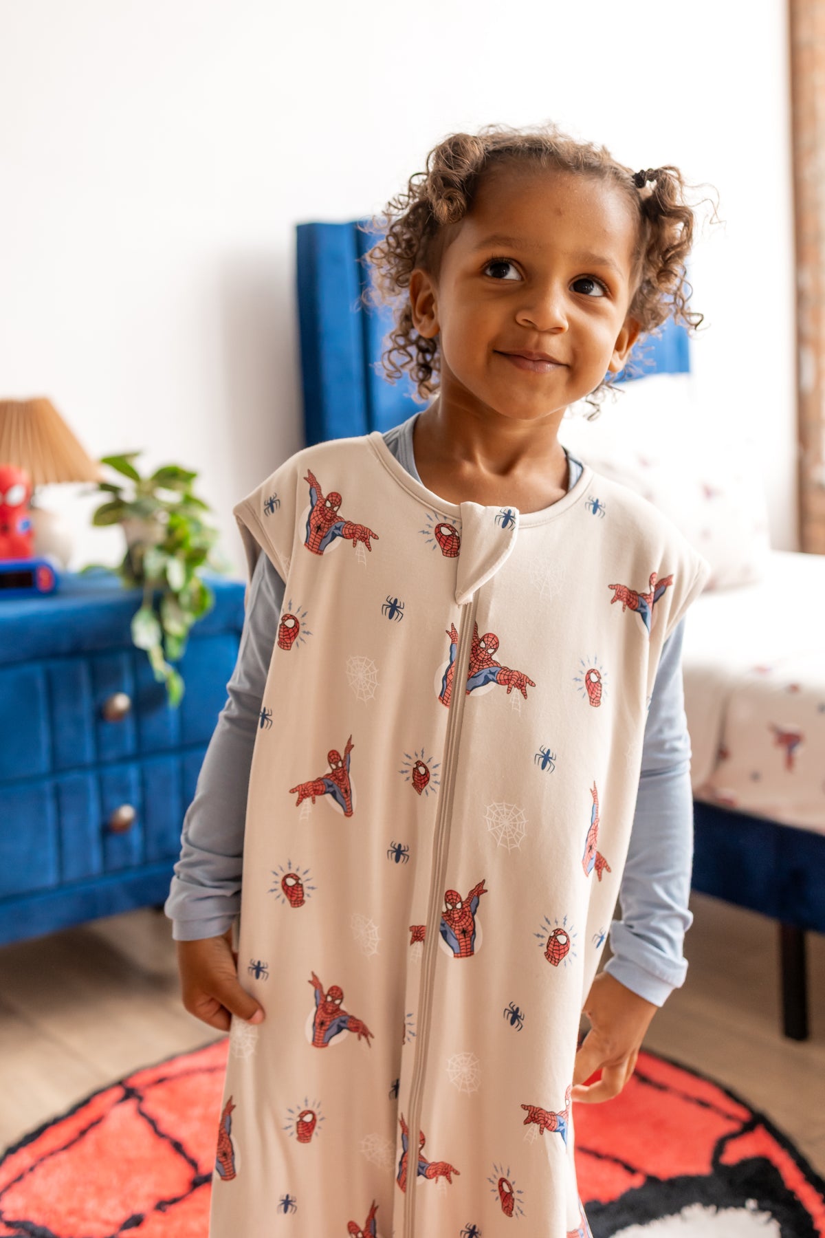 Girl posing on a spider print rug in a room with blue bed and side table wearing oat color Sleep Bag Walker with Spider-Man print 