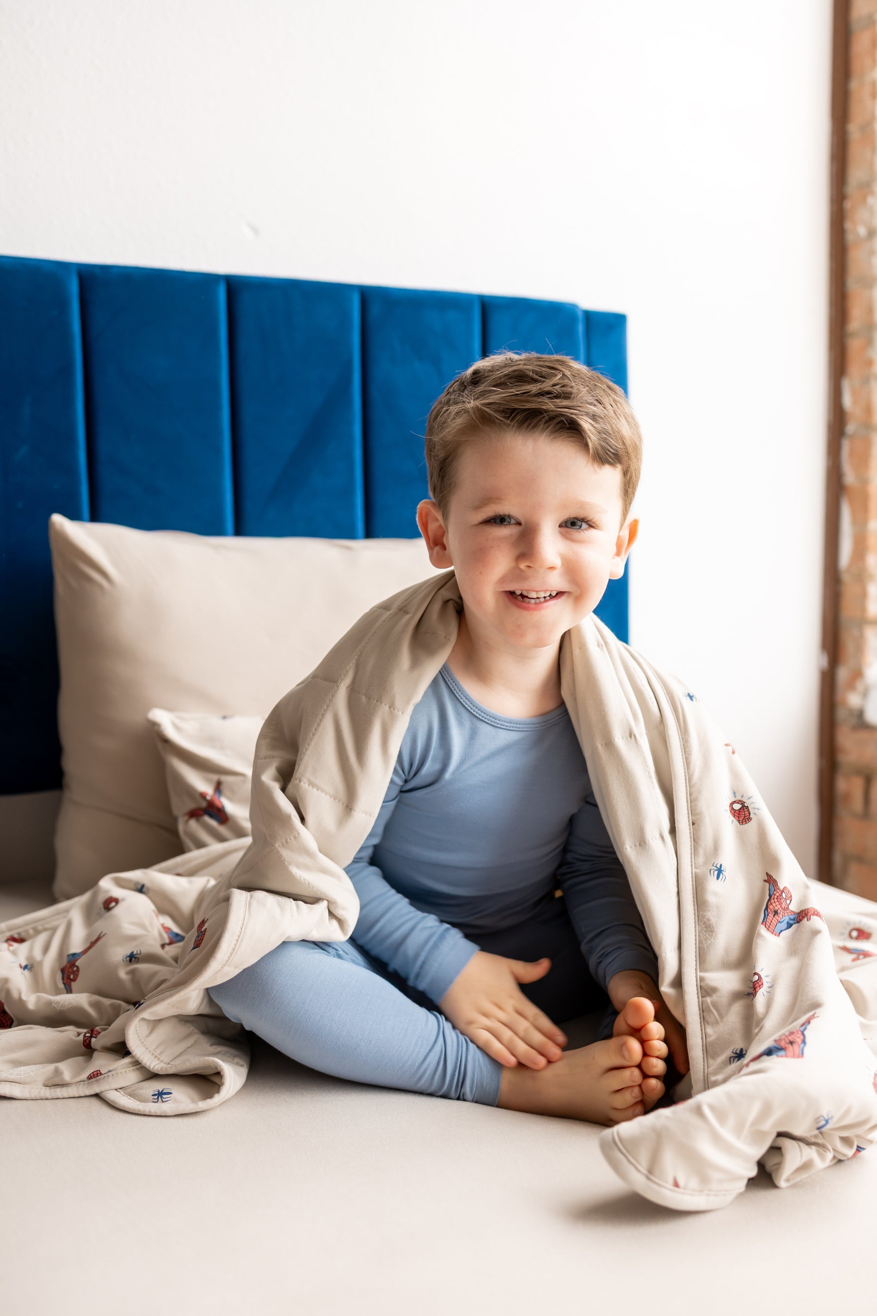 Boy sitting on bed wrapped in oat color Toddler Blanket with Spider-Man print wearing blue pajamas