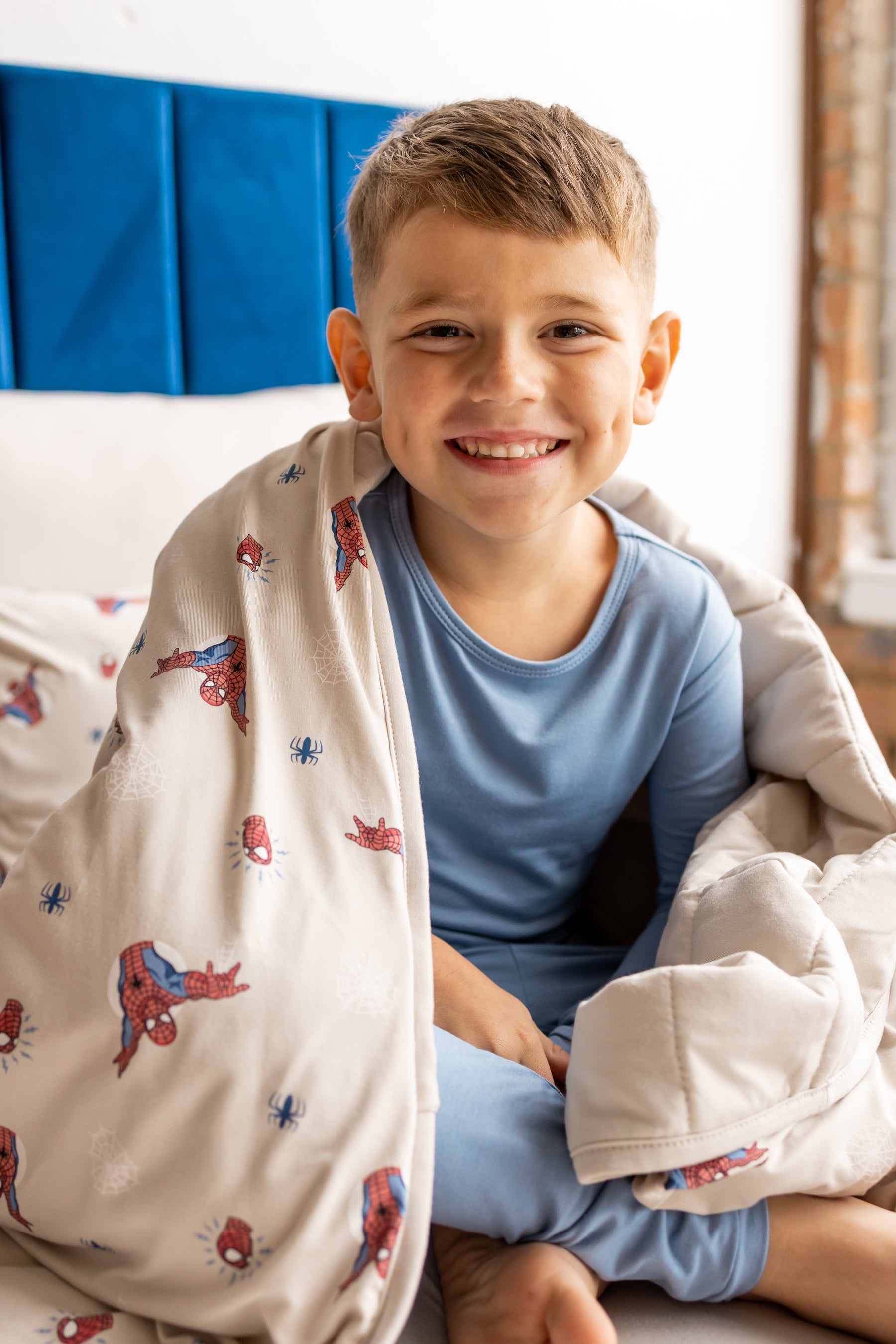 Boy smiling on bed wrapped in oat color Youth Blanket with Spider-Man print wearing blue pajamas