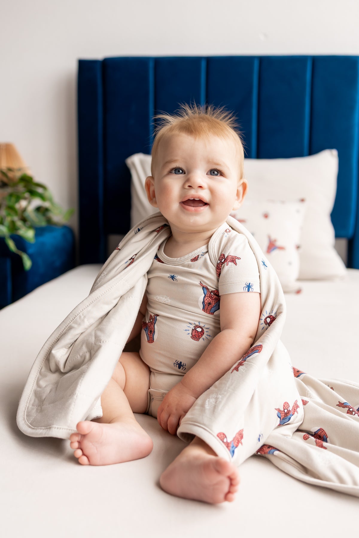 Baby sitting on a beige bed wrapped in an oat color Baby Blanket with Spider-Man print