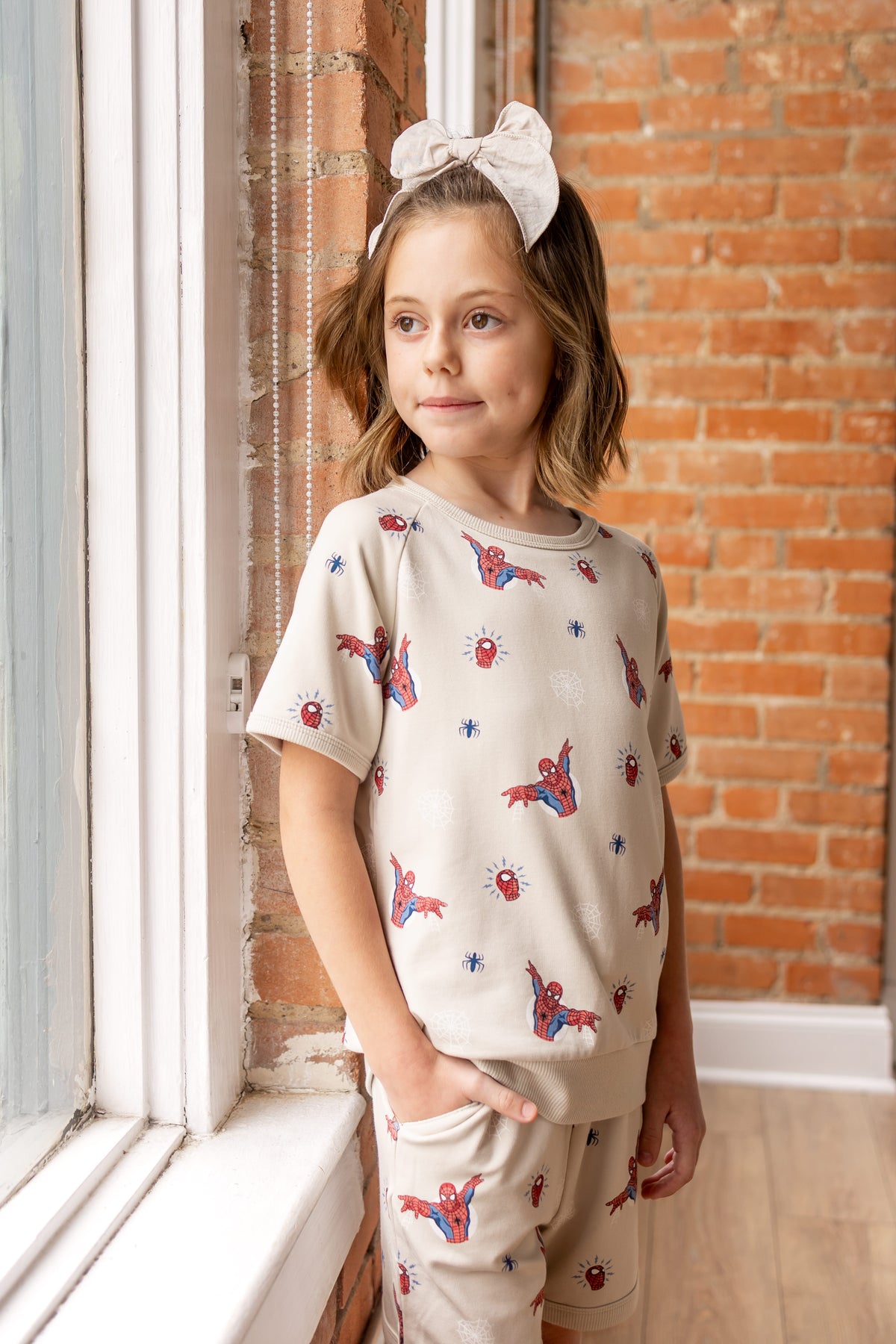 Girl standing in front of a brick wall against a window wearing an oat color Short Sleeve Jogger Set with Spider-Man print and oat color bow on head