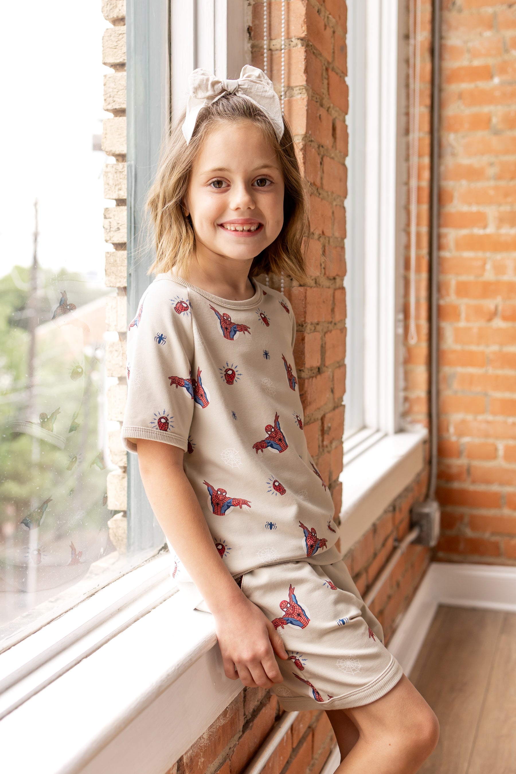 Girl posing in front of a window wearing an oat color Short Sleeve Jogger Set with Spider-Man print and oat color bow on head