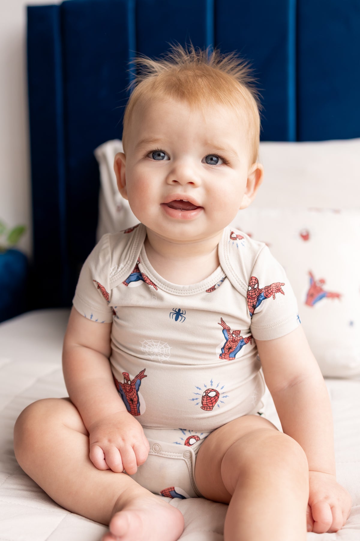 Baby sitting on a bed wearing an oat color Bodysuit in Spider- Man print