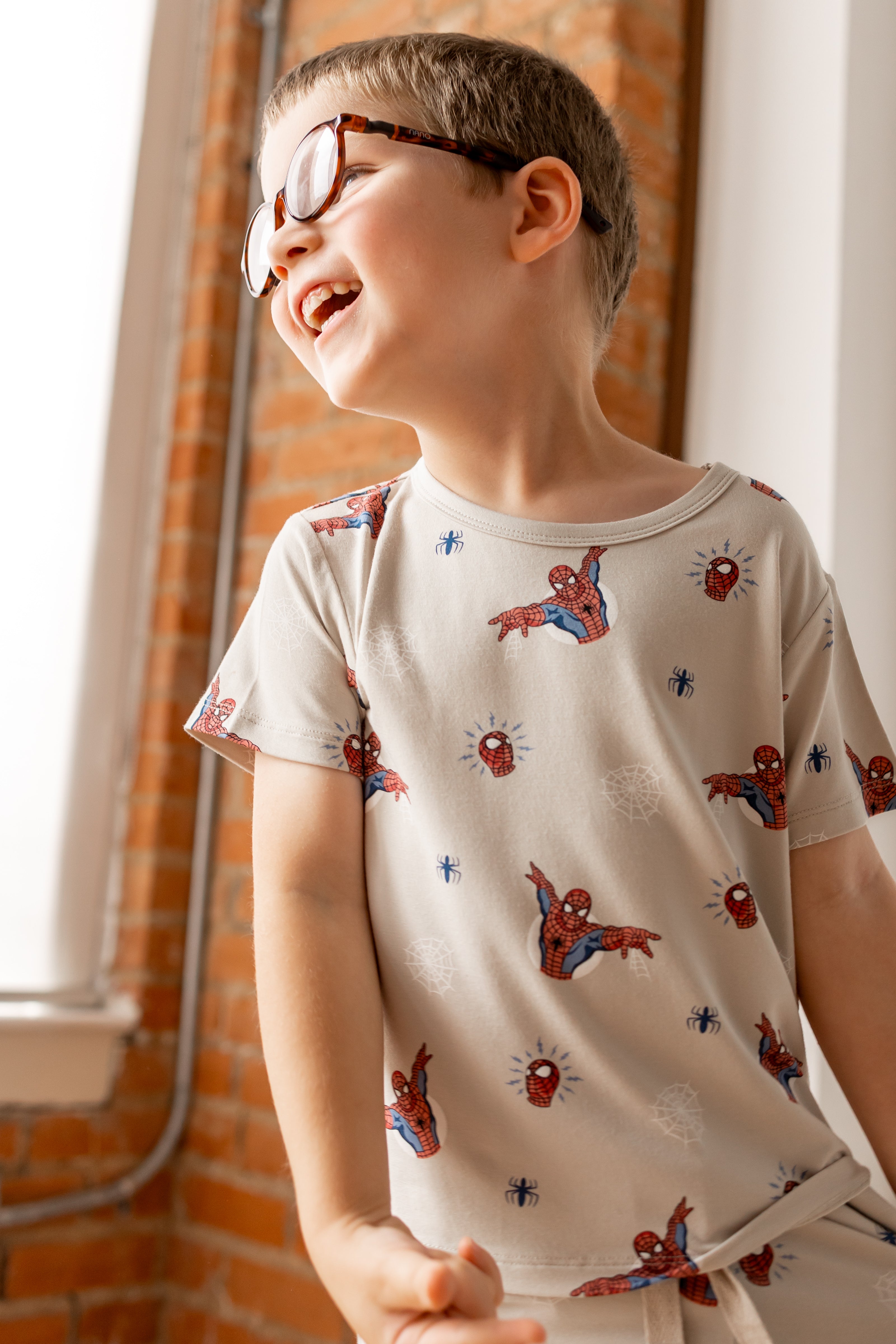 Boy with specs wearing oat color Toddler Basic Tee with Spider-Man print in front of a brick wall 