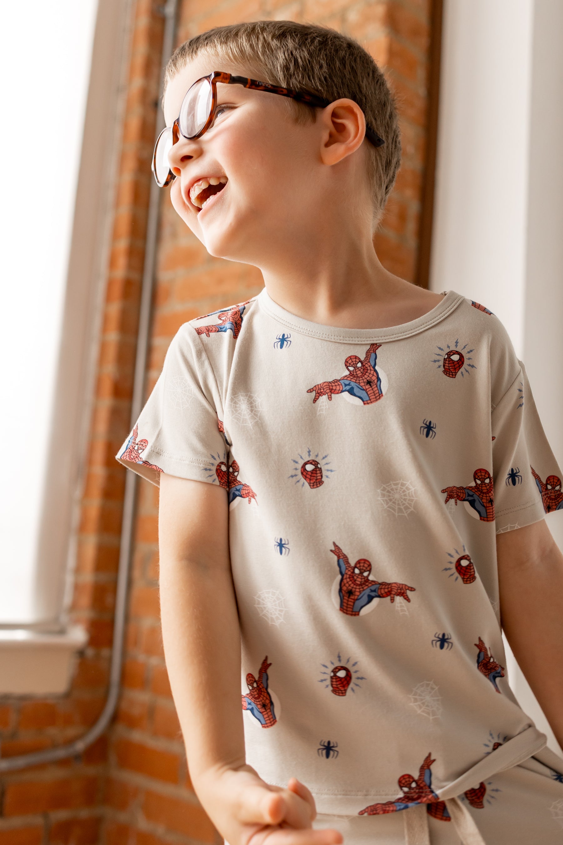 Boy with specs wearing oat color Toddler Basic Tee with Spider-Man print in front of a brick wall 