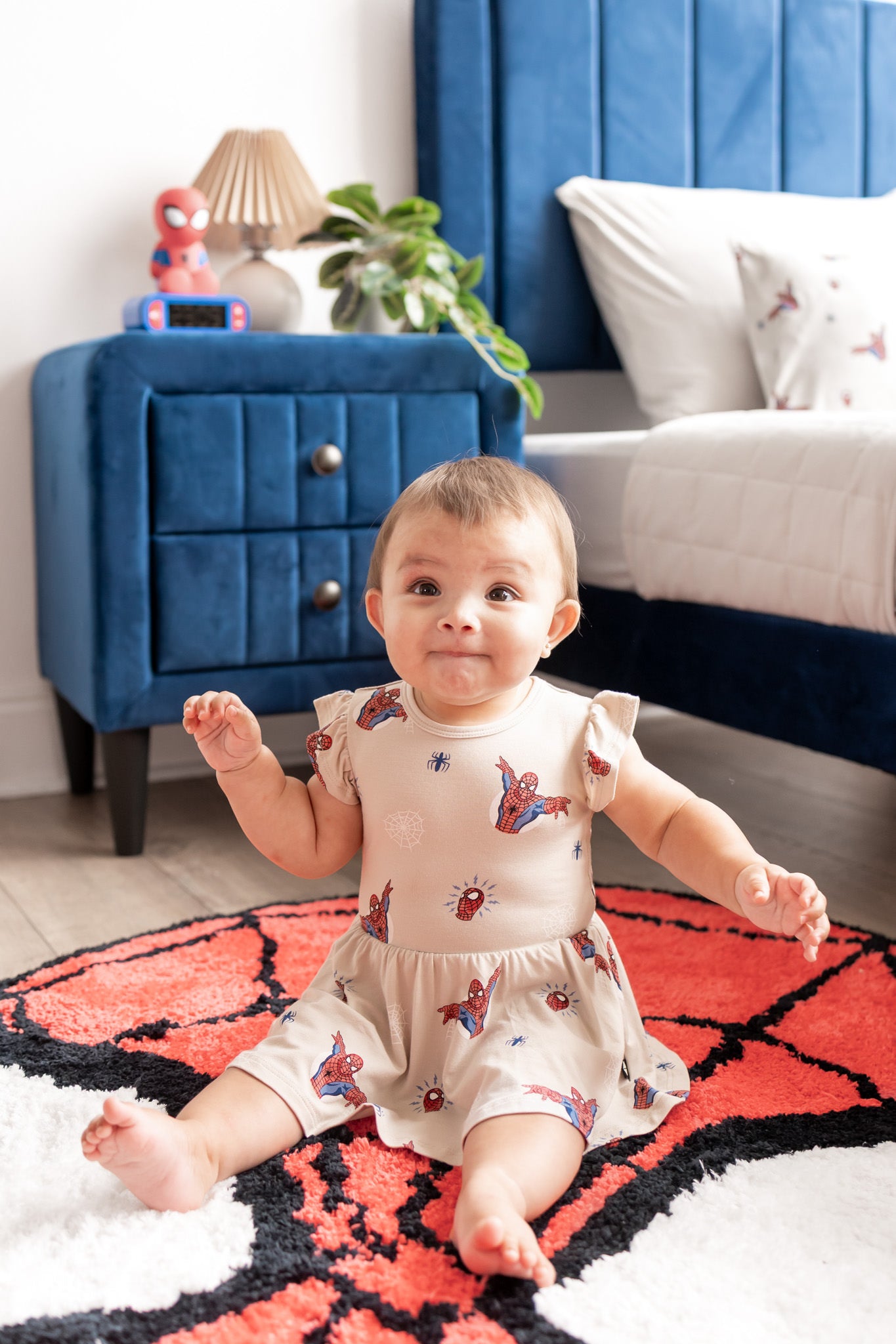 Baby wearing oat color Twirl Bodysuit Dress with Spider-Man print sitting on a spider man print rug in a room with blue bed and side table
