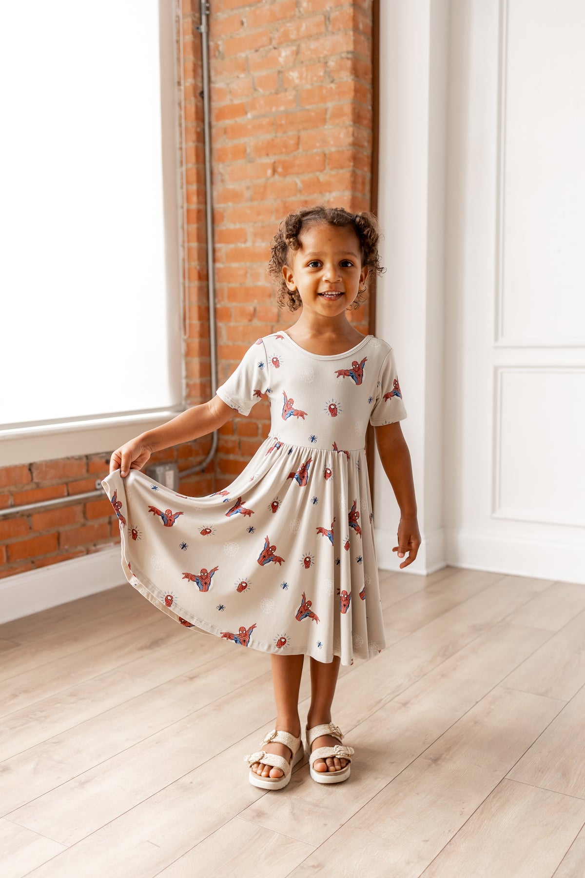 Girl posing in oat color Twirl Dress with Spider-Man print and white sandals in front of a brick wall 