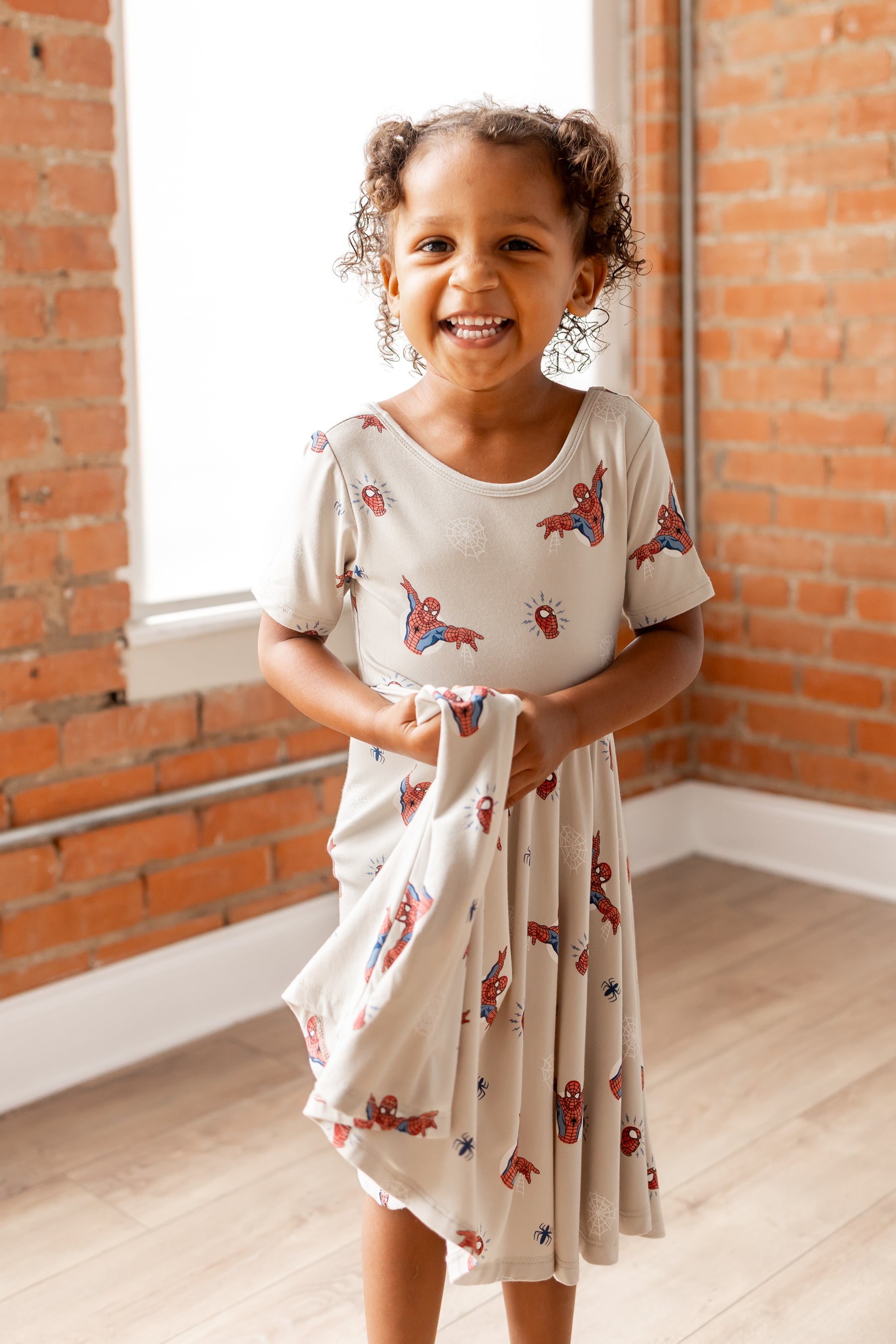 Girl smiling in oat color Twirl Dress with Spider-Man print in front of a brick wall 