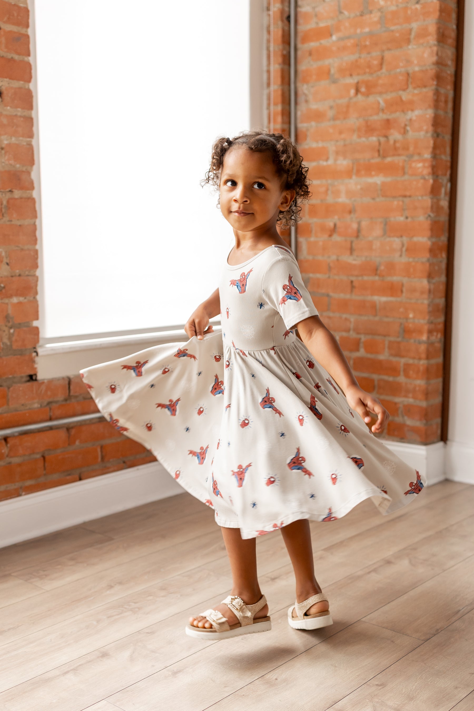Girl twirling wearing oat color Twirl Dress with Spider-Man print and white sandals in front of a brick wall 
