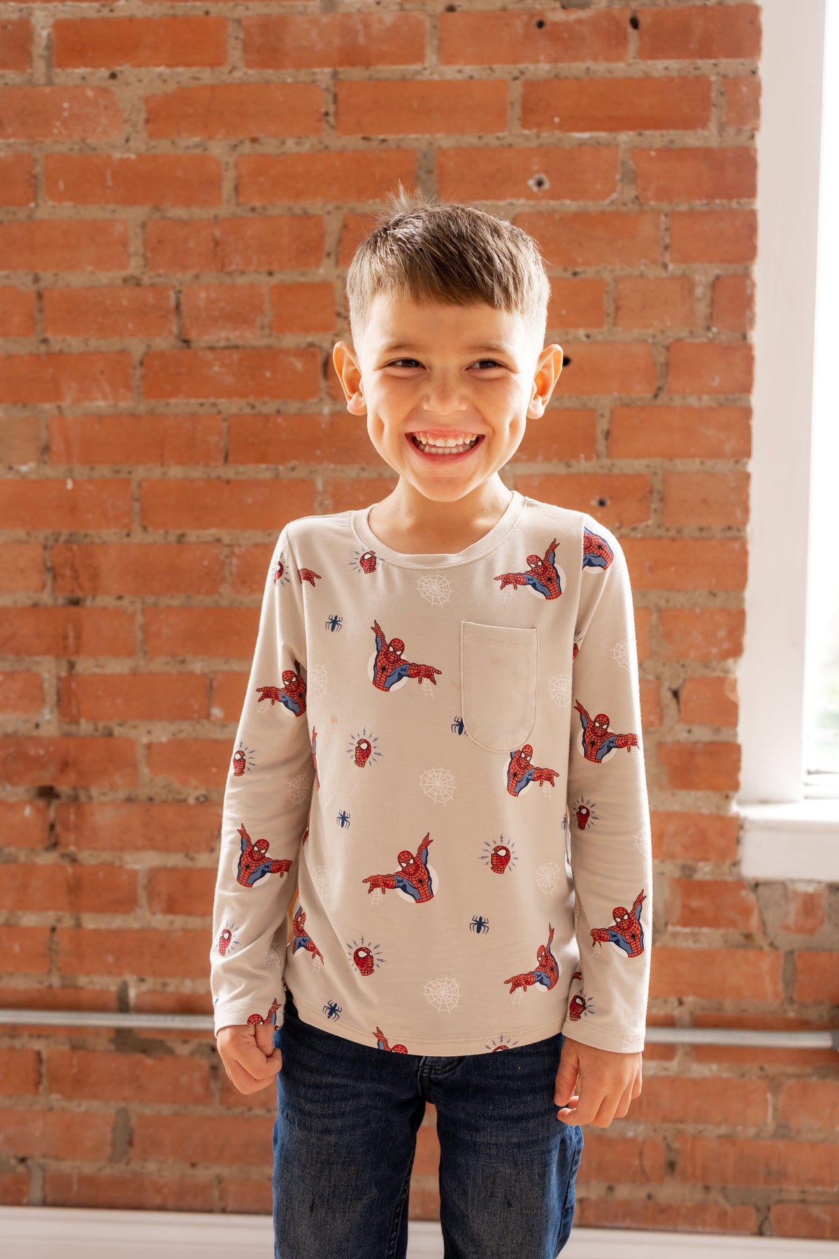 Boy standing in front of a brick wall wearing an oat color Long Sleeve Toddler Crew Neck Tee with Spider-Man print over a jeans