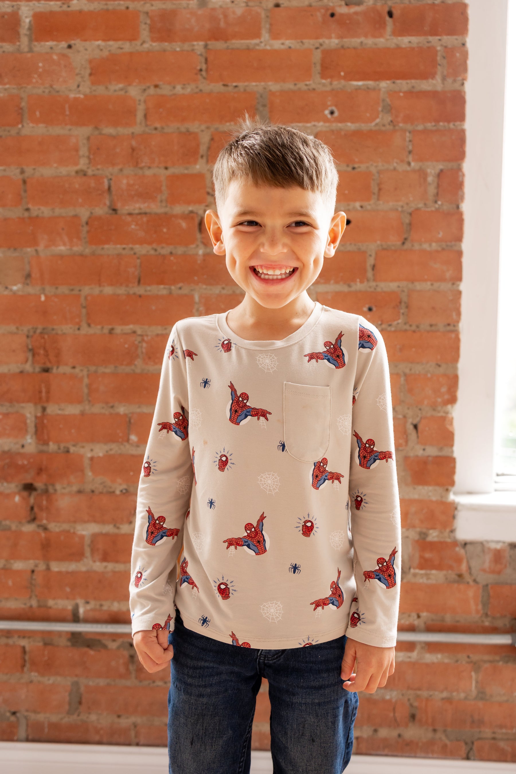 Boy standing in front of a brick wall wearing an oat color Long Sleeve Toddler Crew Neck Tee with Spider-Man print over a jeans