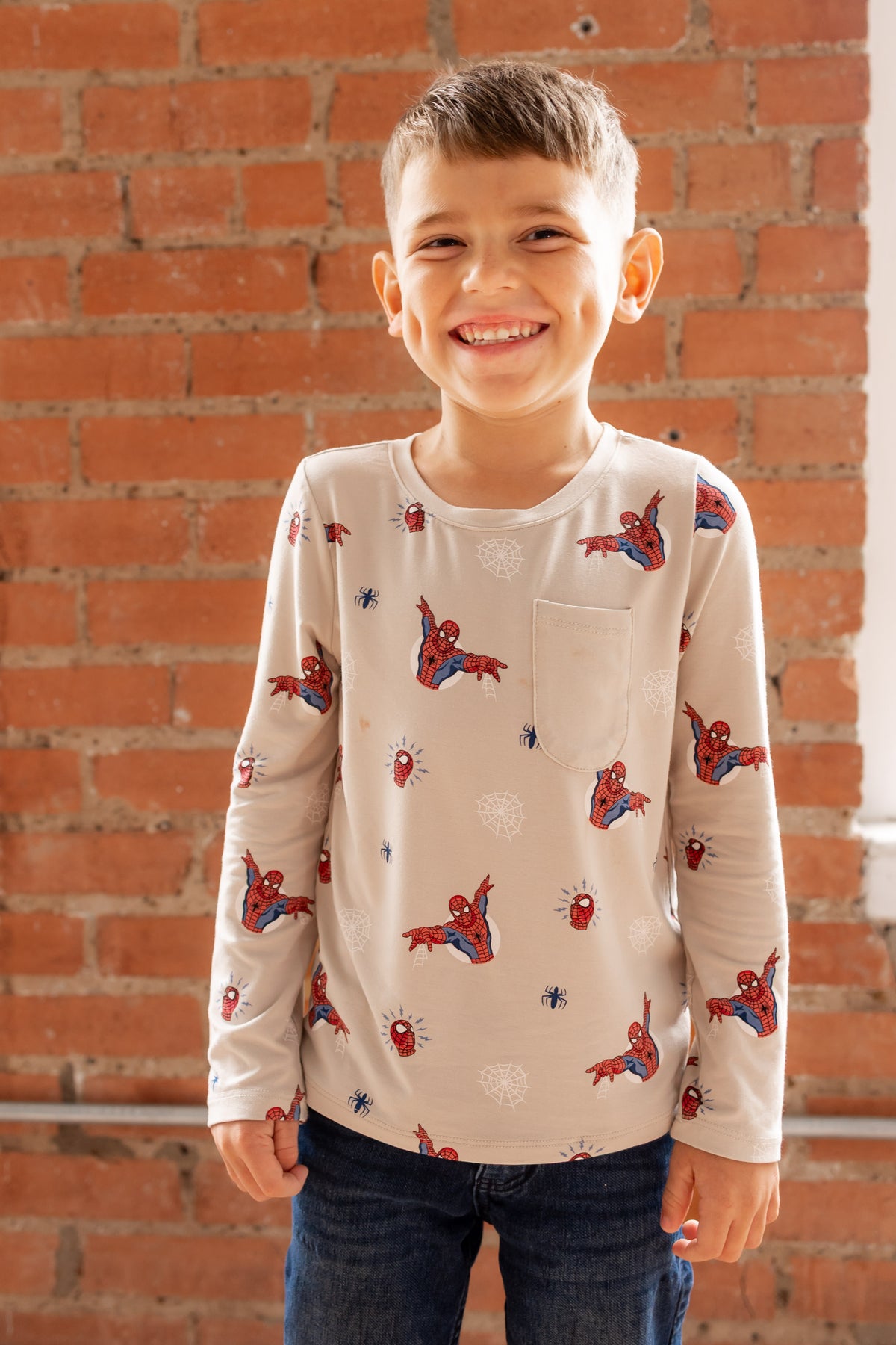 Boy smiling in front of a brick wall wearing an oat color Long Sleeve Toddler Crew Neck Tee with Spider-Man print over a jeans