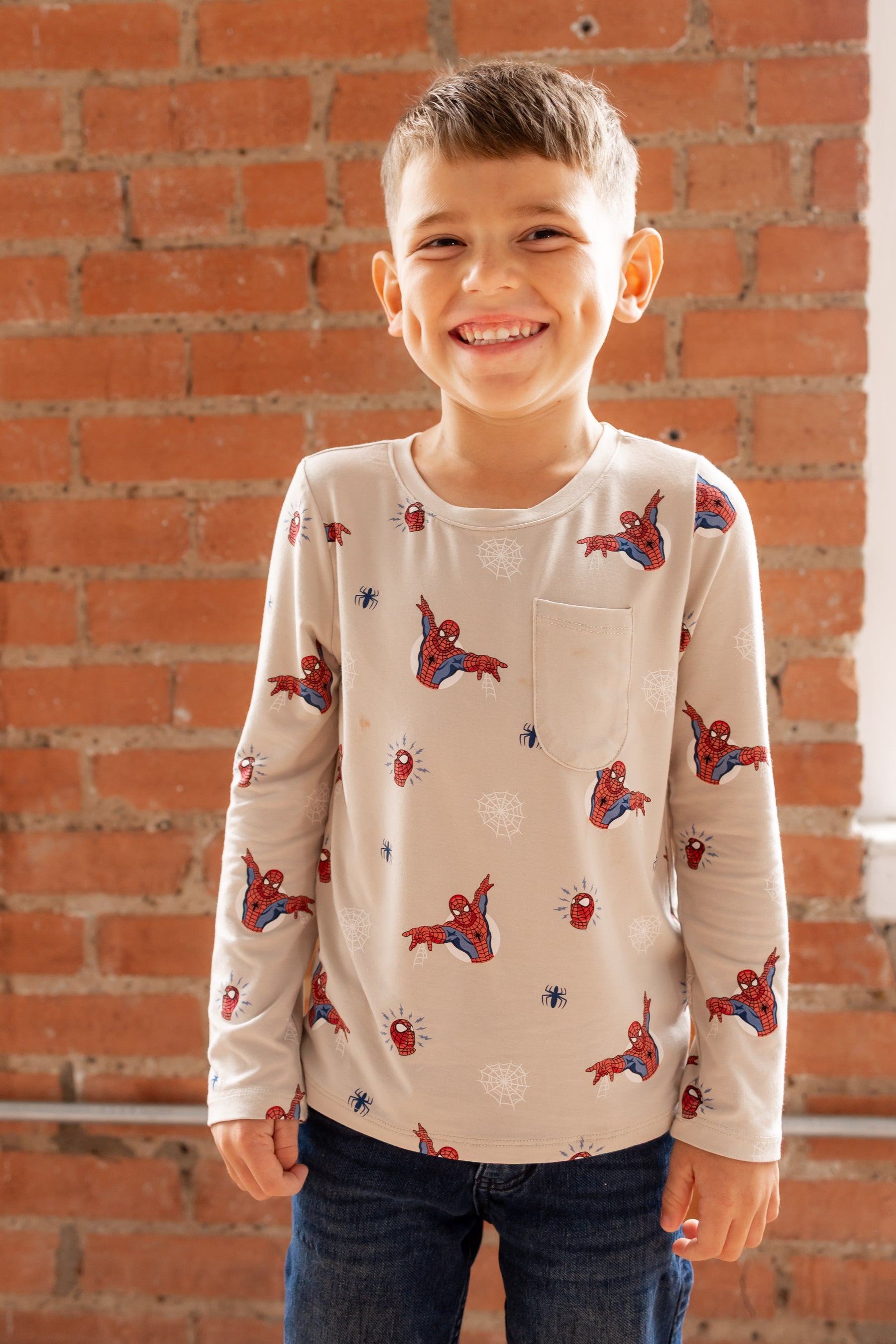 Boy smiling in front of a brick wall wearing an oat color Long Sleeve Toddler Crew Neck Tee with Spider-Man print over a jeans