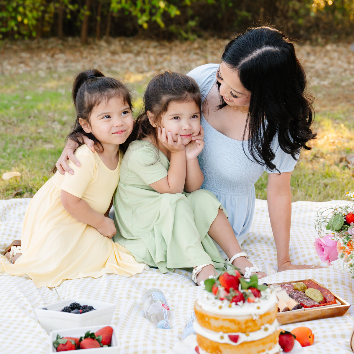 Two daughters wearing the Twirl Dress in Chamomile and Basil sitting having a picnic with their mom wearing the Women's Smocked Dress in Breeze