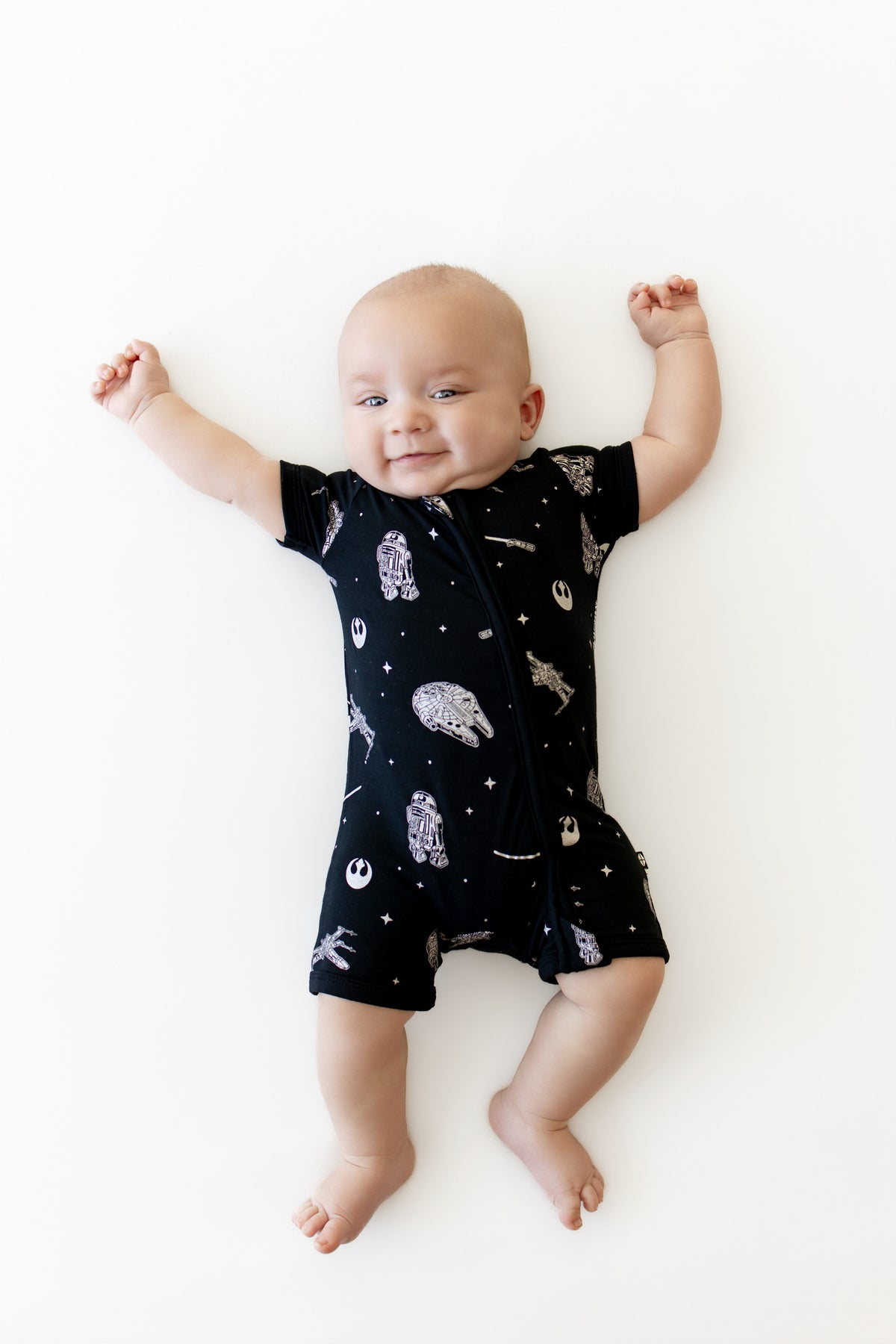 Infant stretched out on a white surface wearing the Zipper Shortall Romper in STAR WARS™