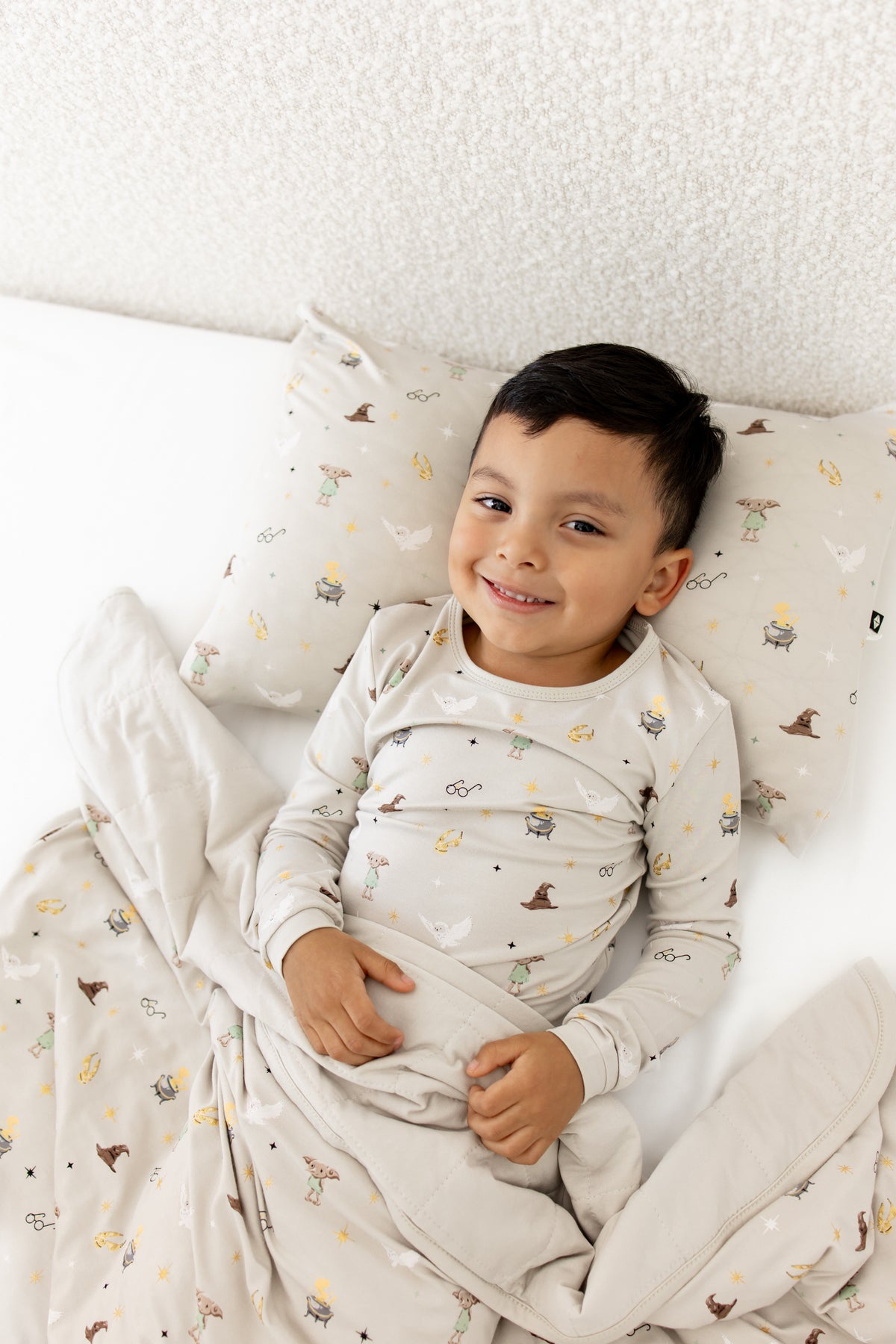 Child lying under a beige Toddler blanket and a pillowcase with Harry Potter prints on it wearing a similar print shirt on a white sheet.