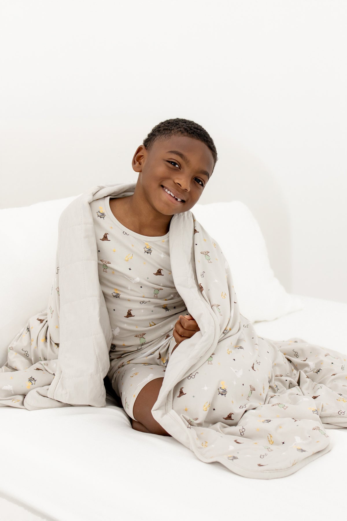 Child wrapped in a beige Youth blanket with Harry Potter prints wearing a similar print short sleeve pajama set sitting on a white bed sheet.