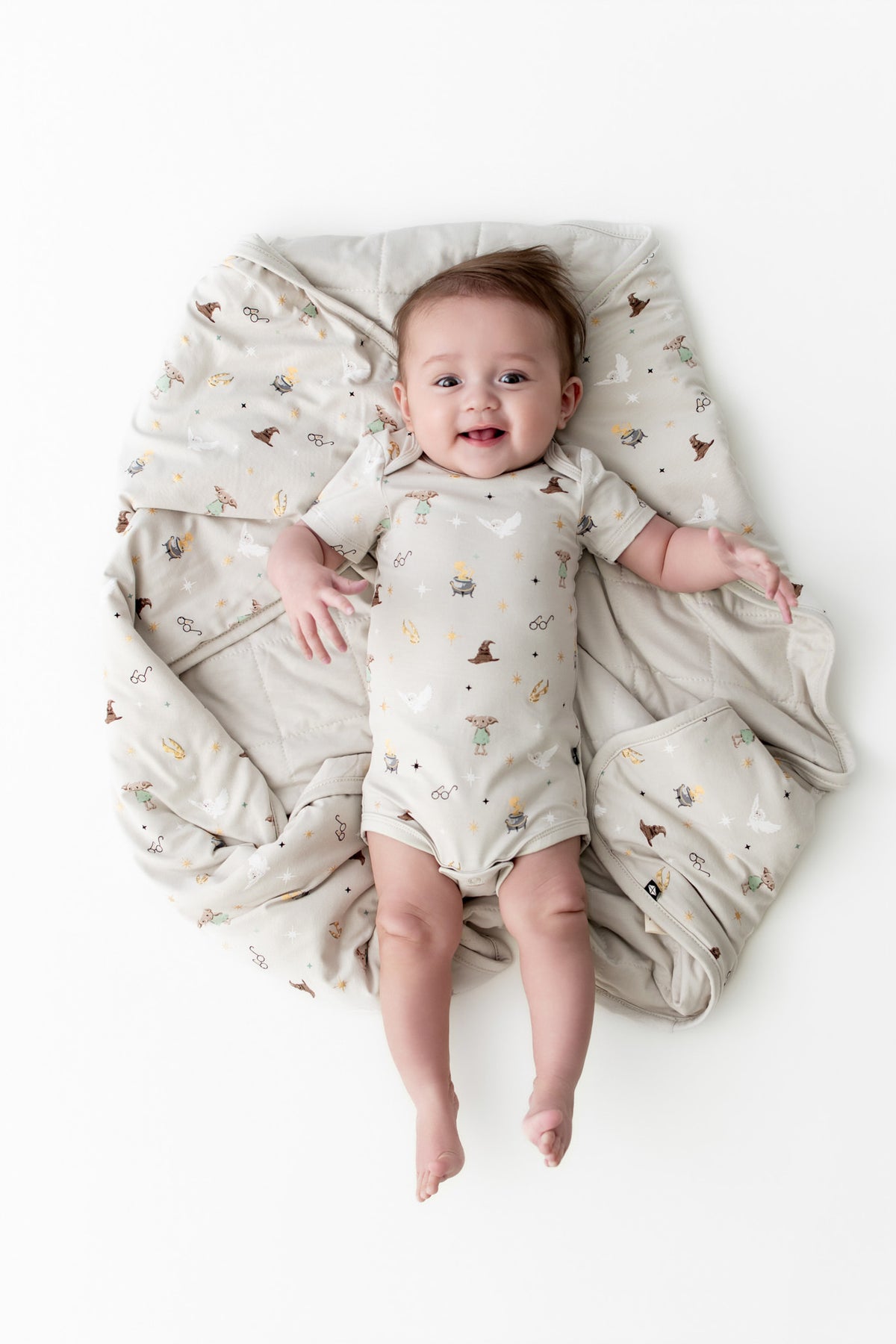 Baby wrapped in a beige baby blanket with Harry Potter patterns wearing a similar bodysuit on a white background
