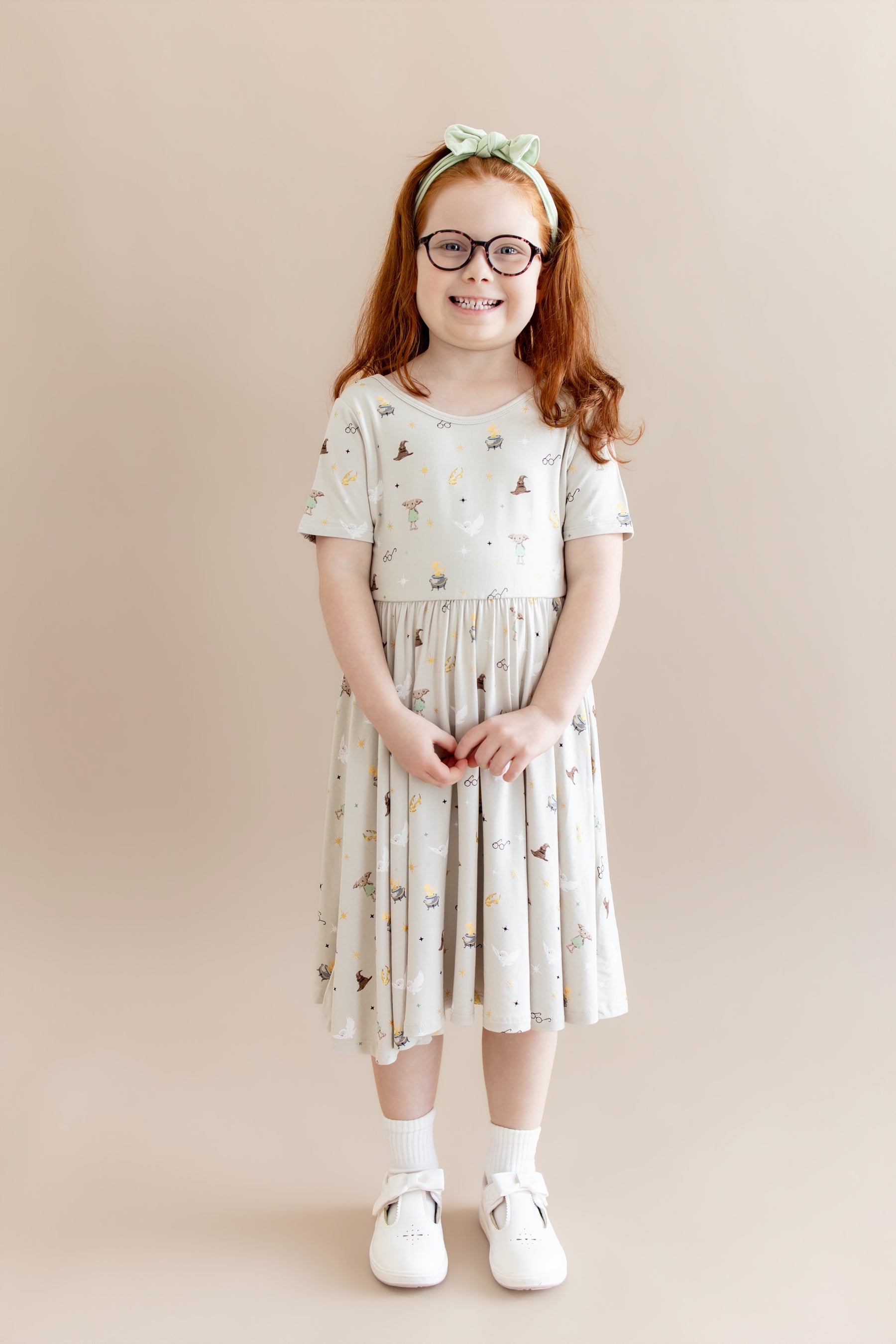 Child wearing a Harry Potter patterned twirl dress with a green headband and white socks and shoes on a beige background