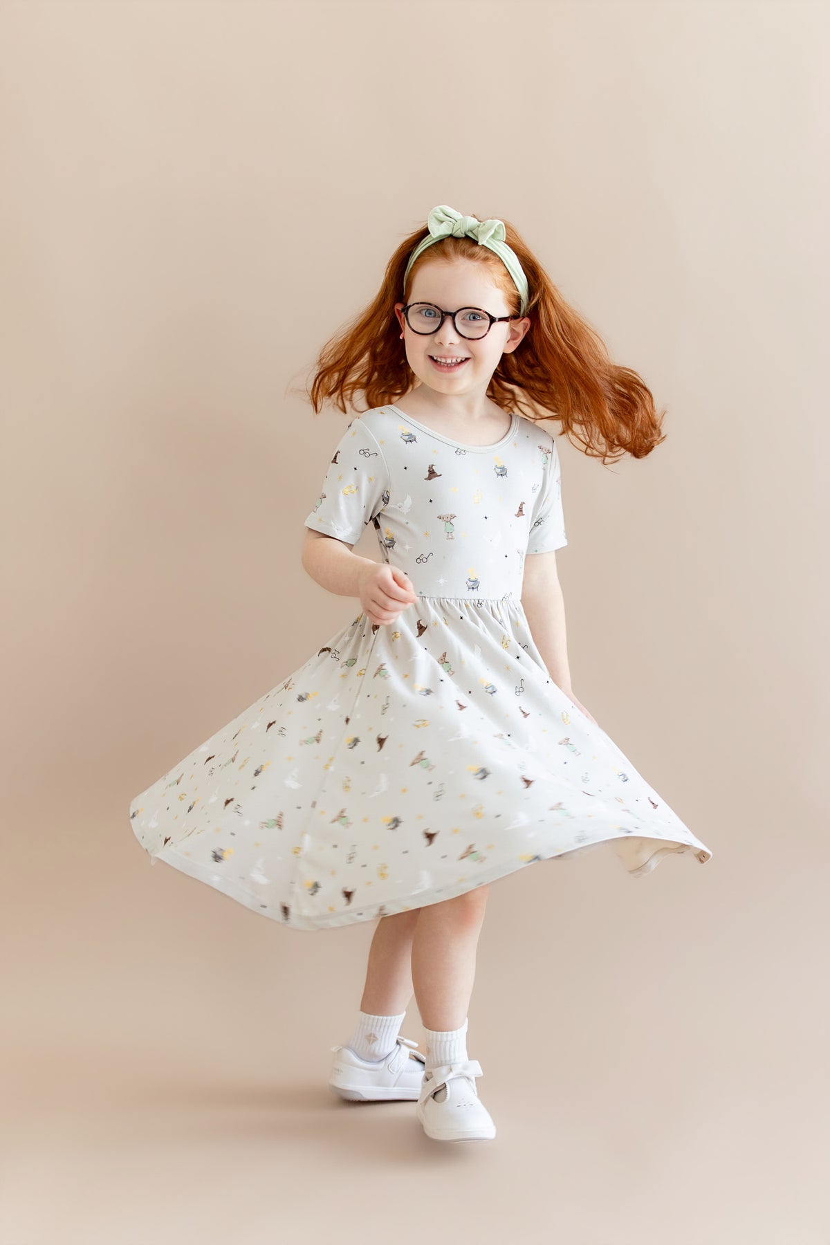 Child twirling in a Harry Potter patterned twirl dress with a green headband and white socks and shoes on a beige background