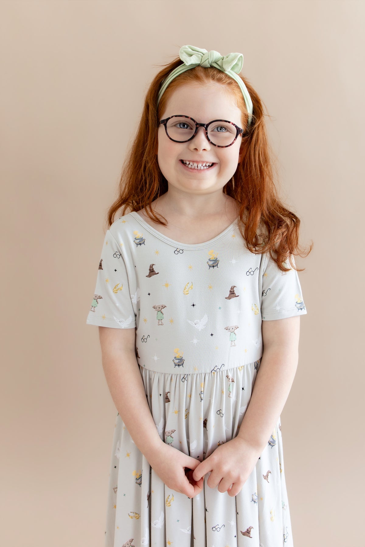 Child wearing a cream Twirl dress with Harry Potter prints on it with a green headband on a beige background