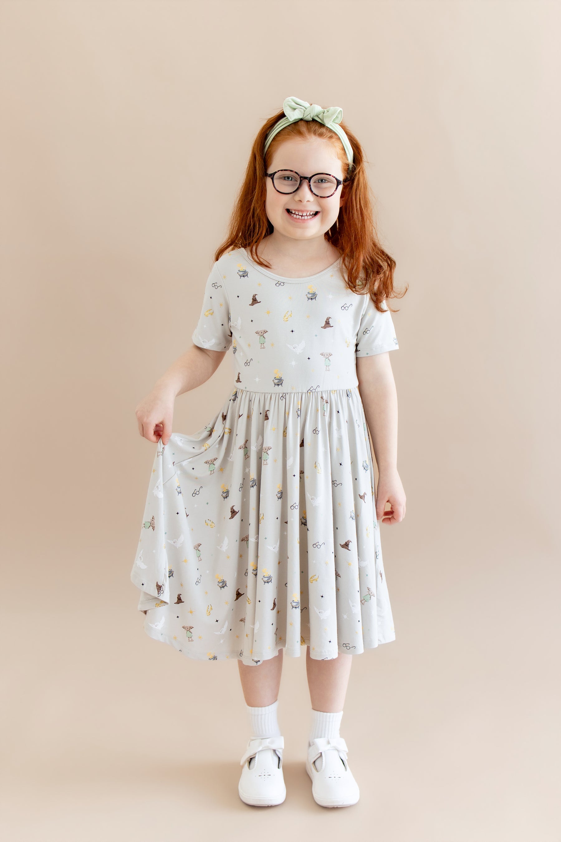 Child wearing a cream Twirl dress with Harry Potter prints with white socks and shoes and green headband on a beige background