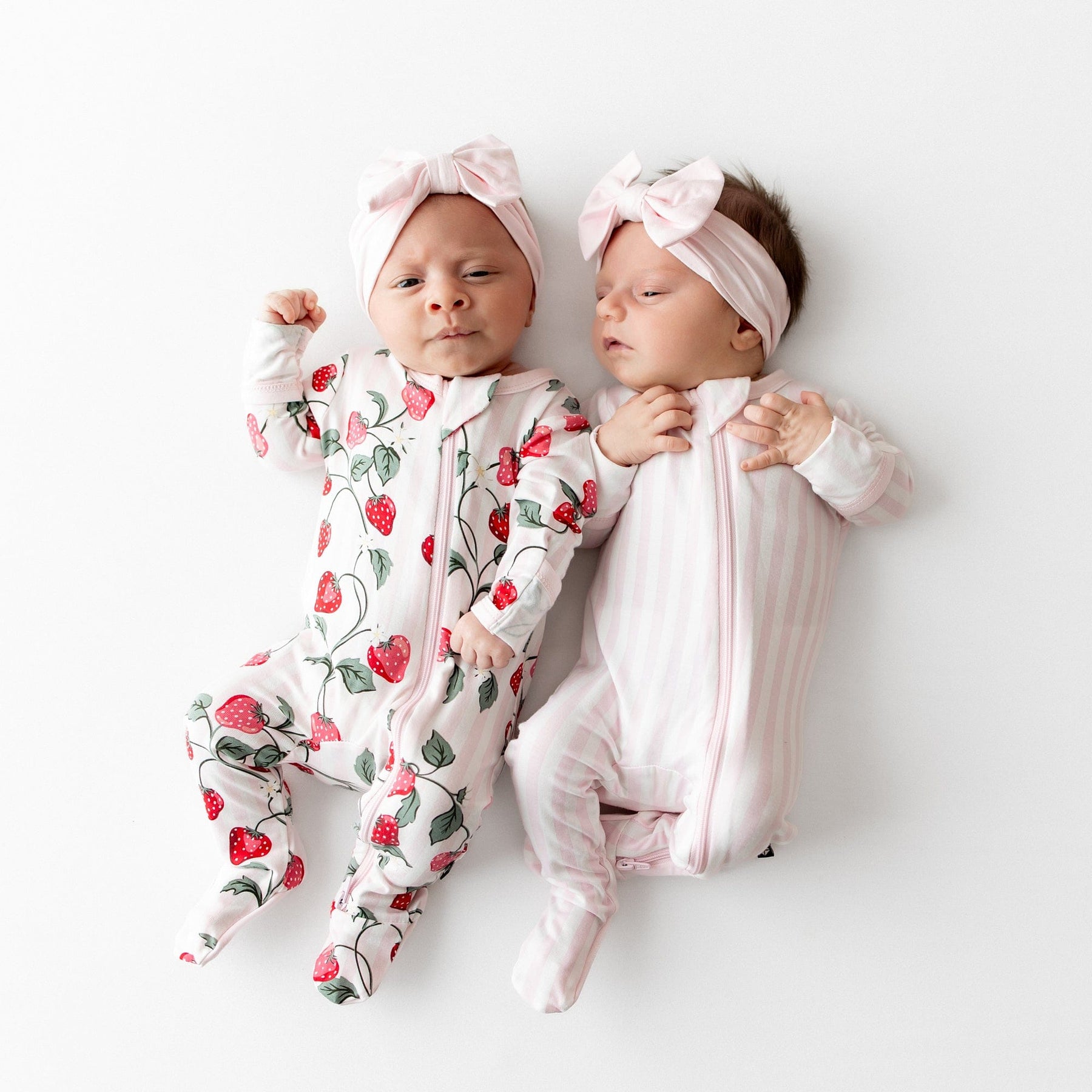 Two babies wearing strawberry-patterned and pink striped outfits with headbands on a white background