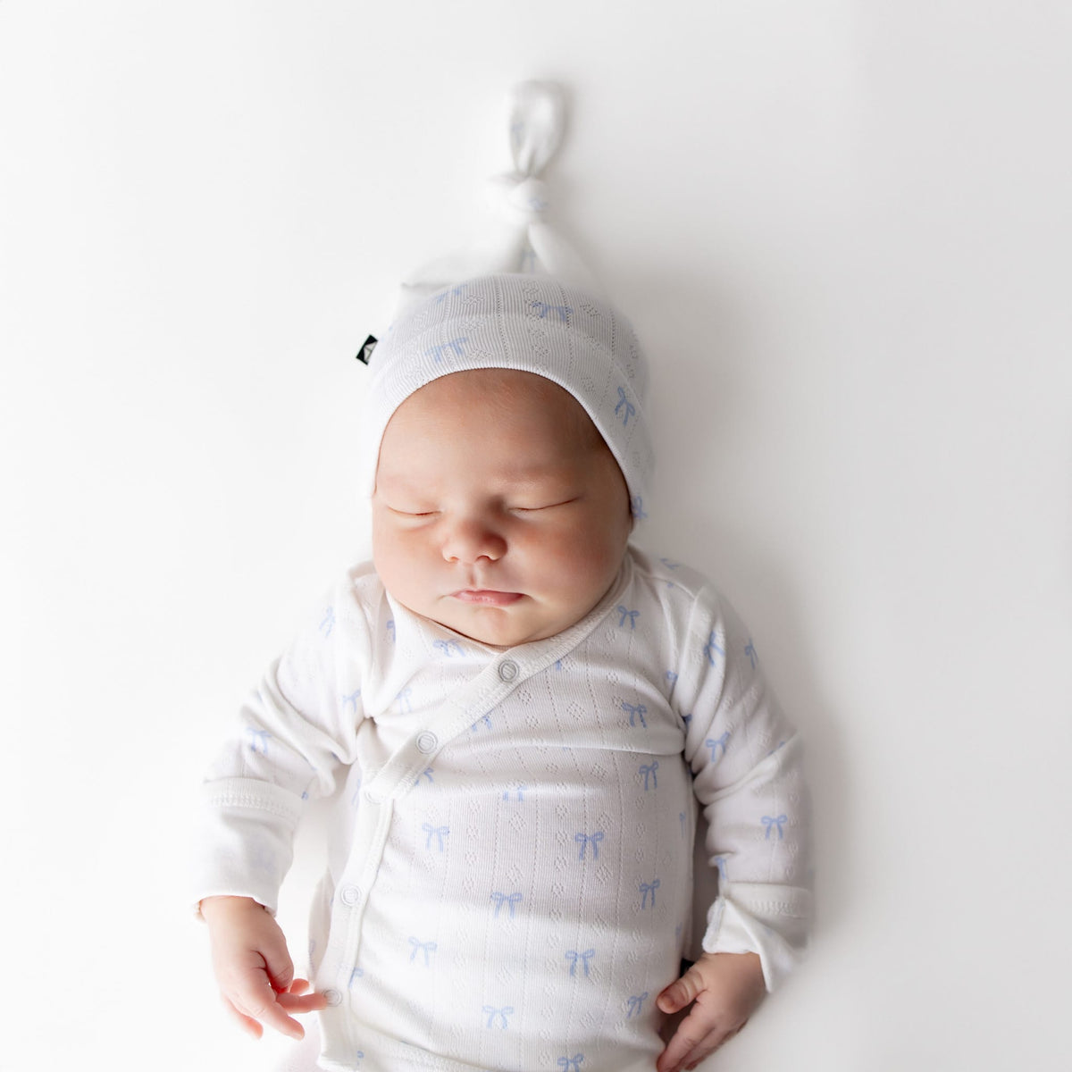 Newborn baby wearing a white outfit with blue patterns and a matching hat, lying on a white background.