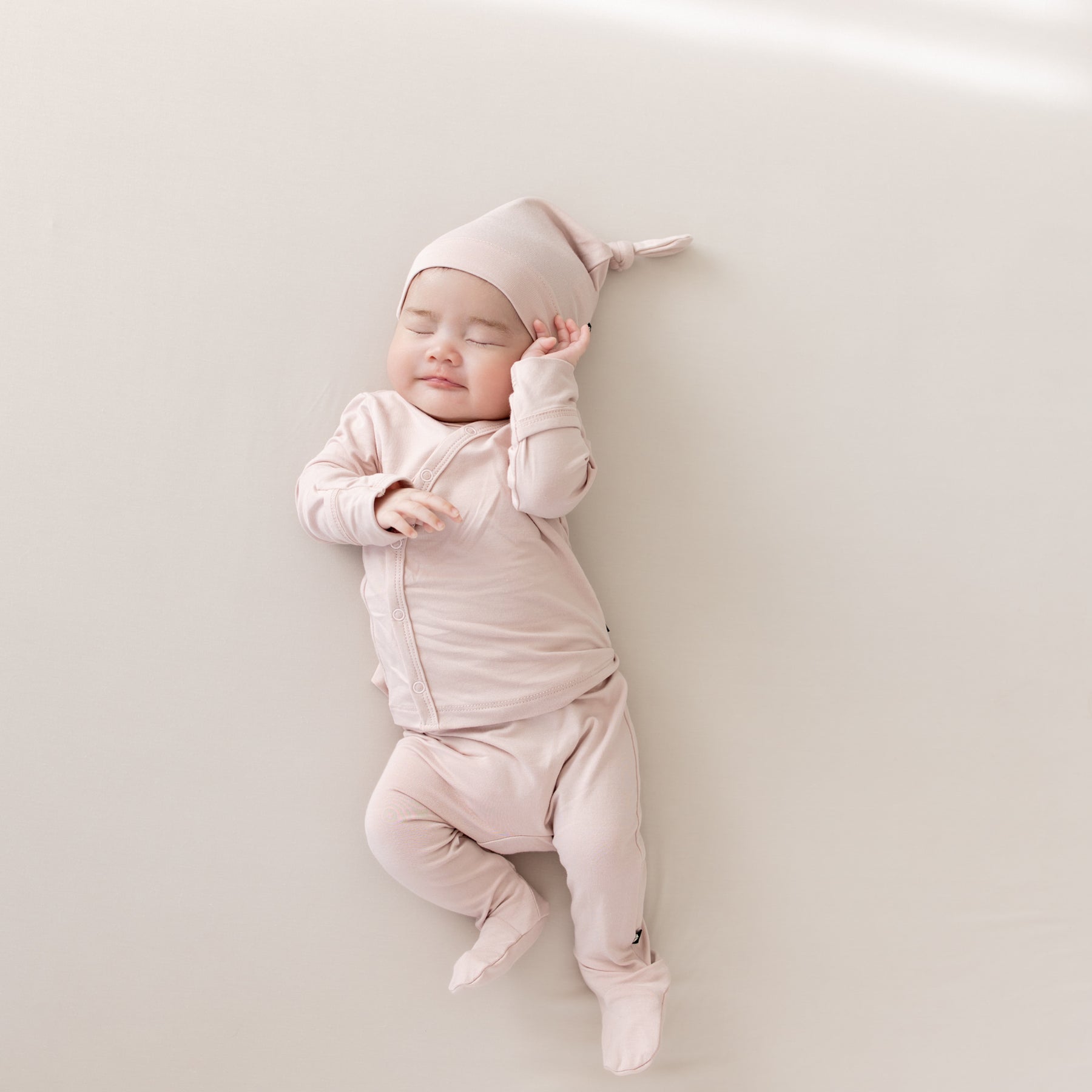Baby laying on an Oat surface while wearing the Bamboo Take Me Home Set with Cap in Blush 