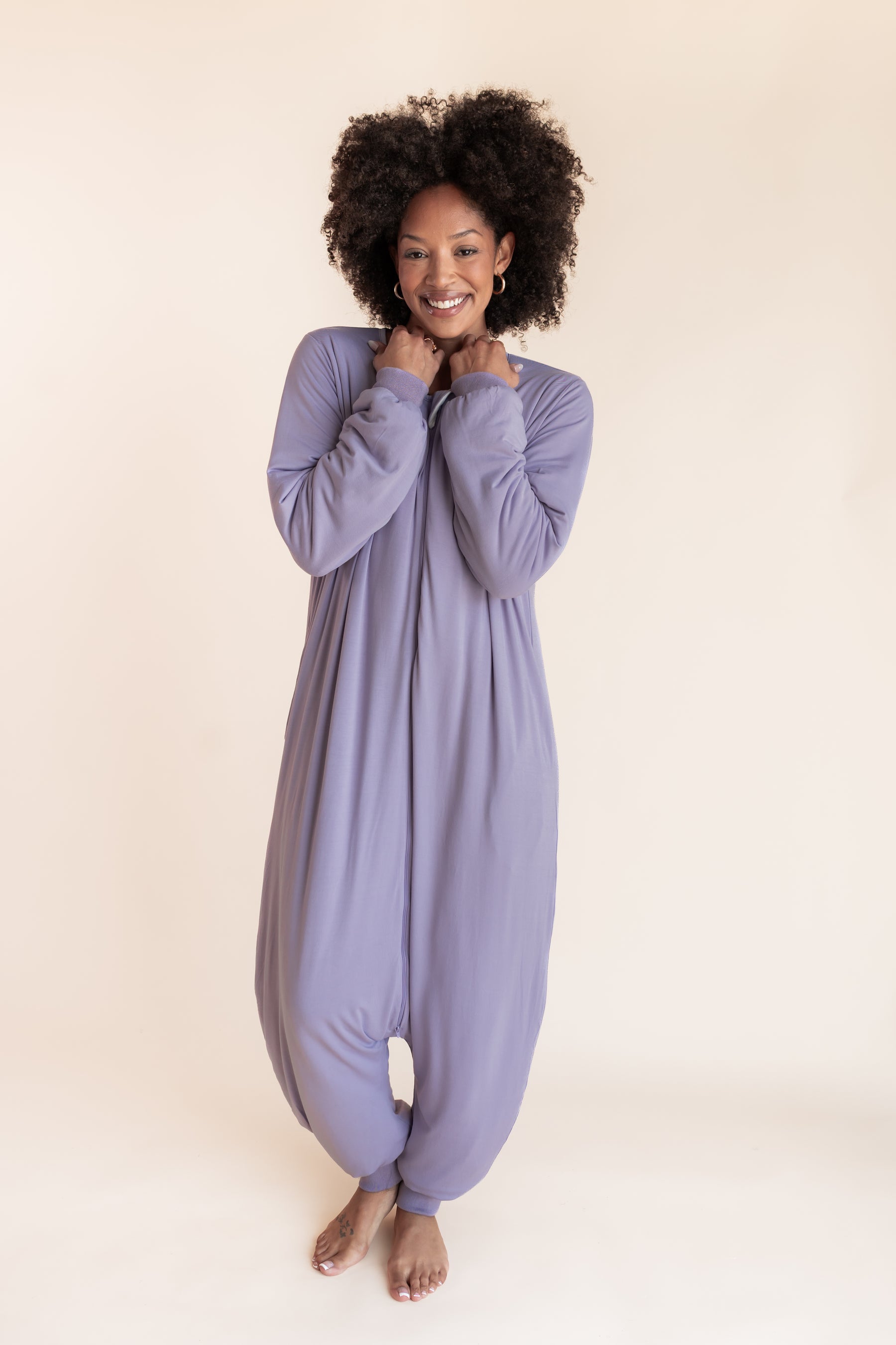 A woman with curly black hair wearing a lavender-purple long-sleeve harem-style sleep sack, standing against a solid light beige background.