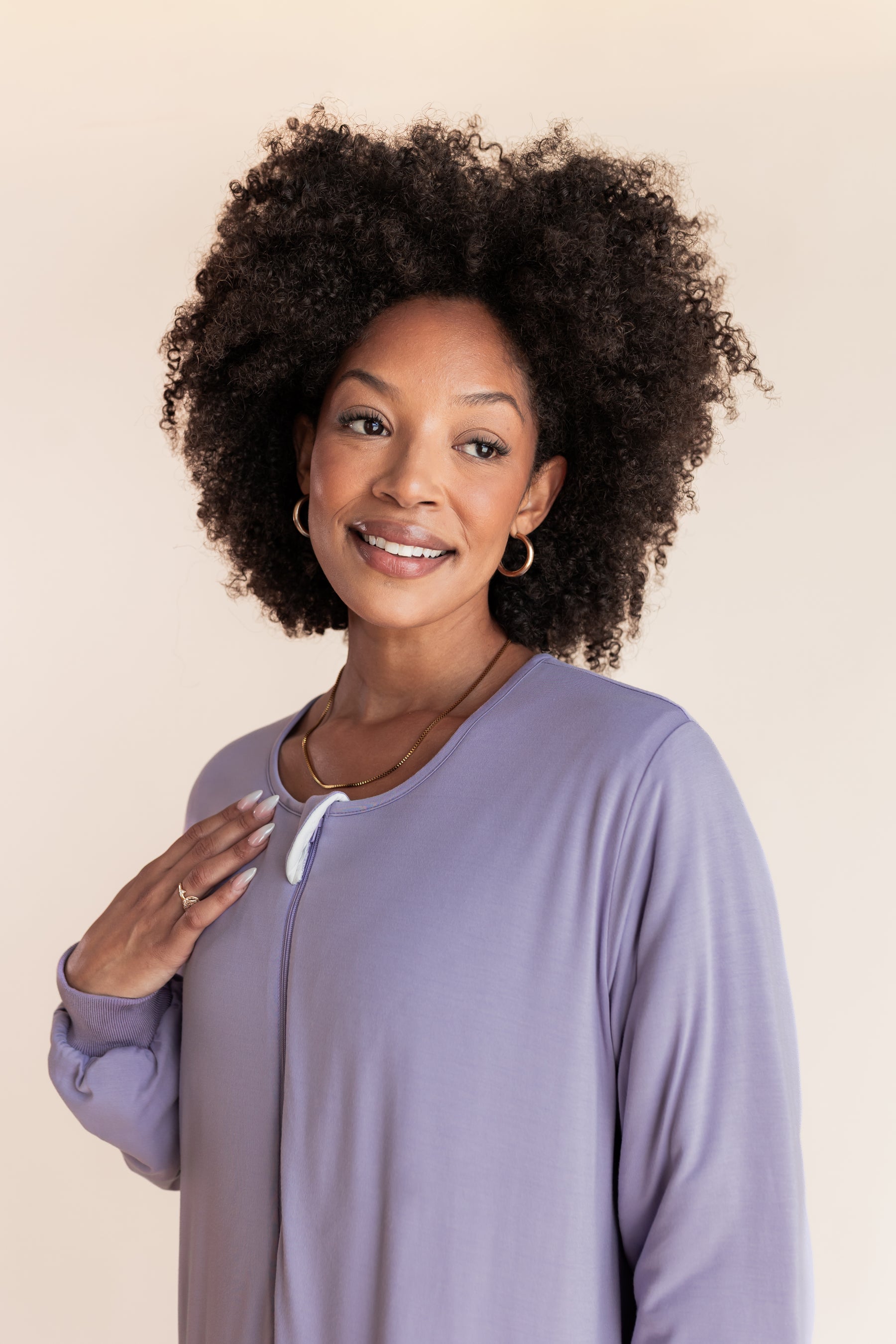 A smiling woman with voluminous dark curly hair wearing a lavender long-sleeve sleep sack with a white drawstring, photographed against a solid light beige background.