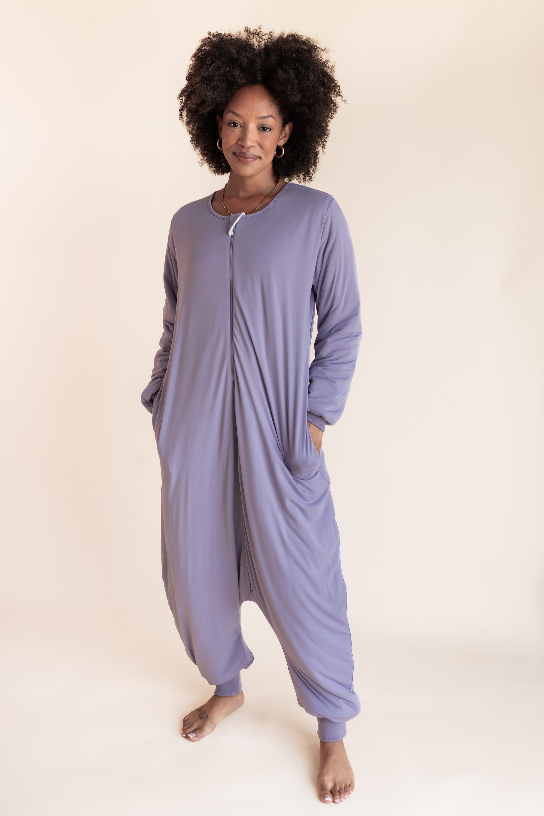 A woman modeling a lavender-purple long-sleeve zip-up harem onesie against a solid cream studio background.