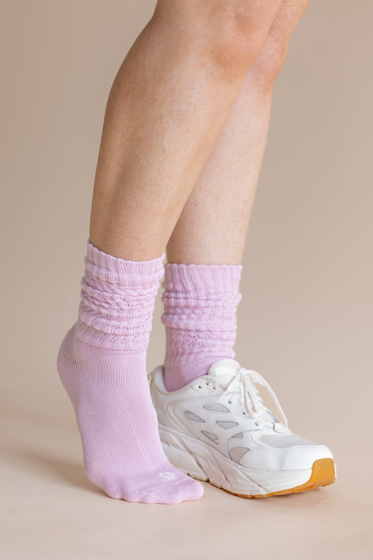 A close-up of a person's feet wearing light purple ribbed crew socks and a white athletic sneaker against a solid beige background.