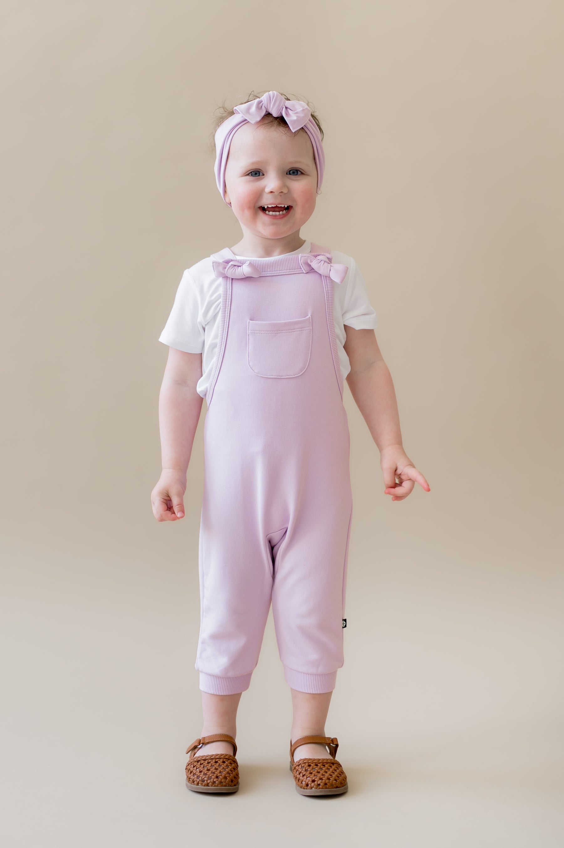 Young girl standing in front of a beige background wearing the Overall in Thistle