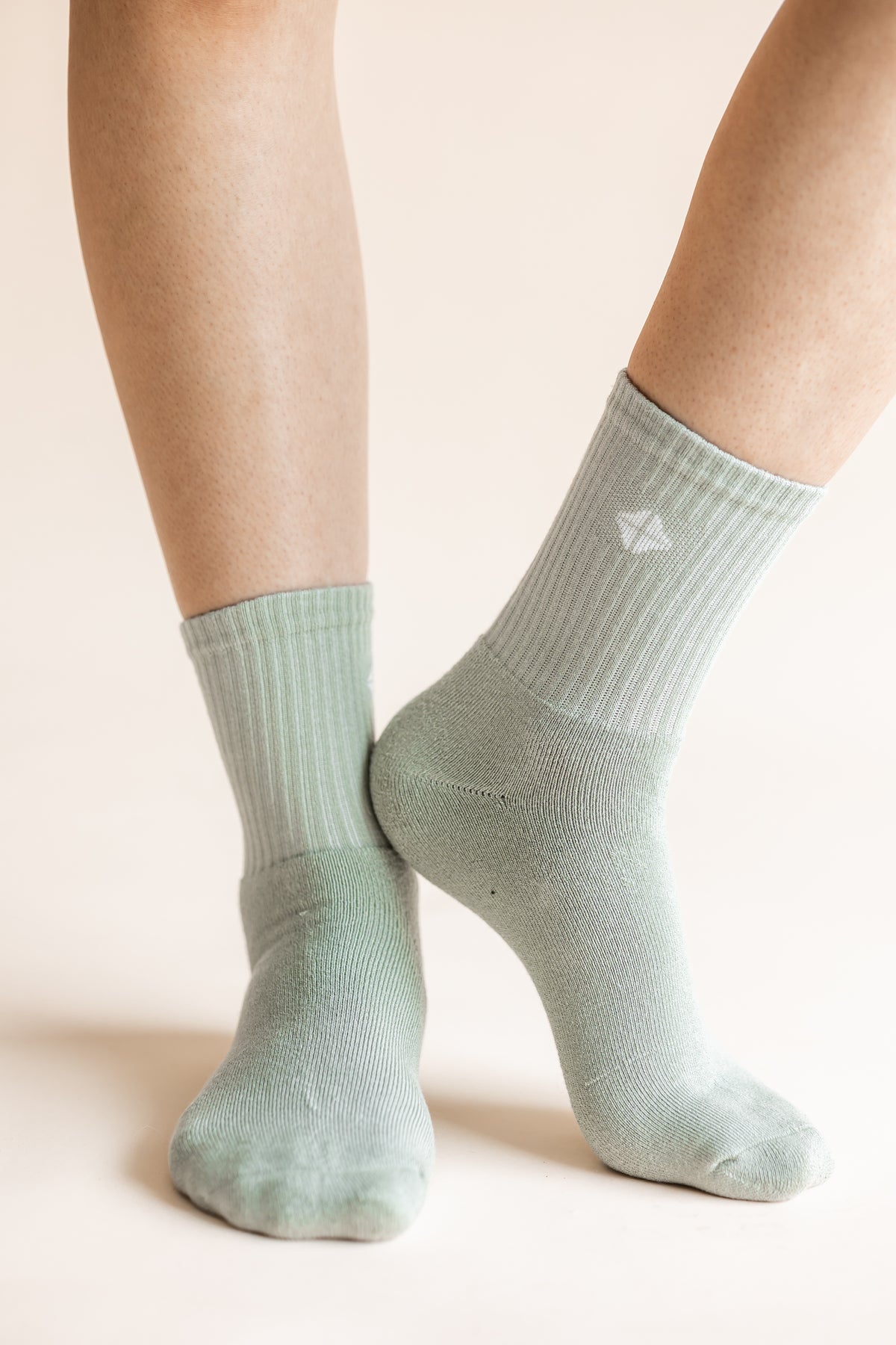 A close-up of a person's feet wearing ribbed crew socks in a pale sage green color, with a small white geometric logo on the side of the right sock.
