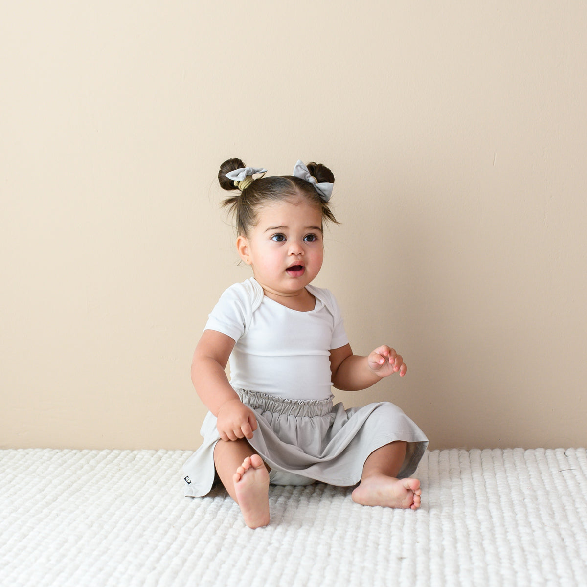 Toddler sitting on the floor while wearing a Toddler Skort in Oat, Short Sleeve Bodysuit in Cloud and pigtails with Bows in Oat