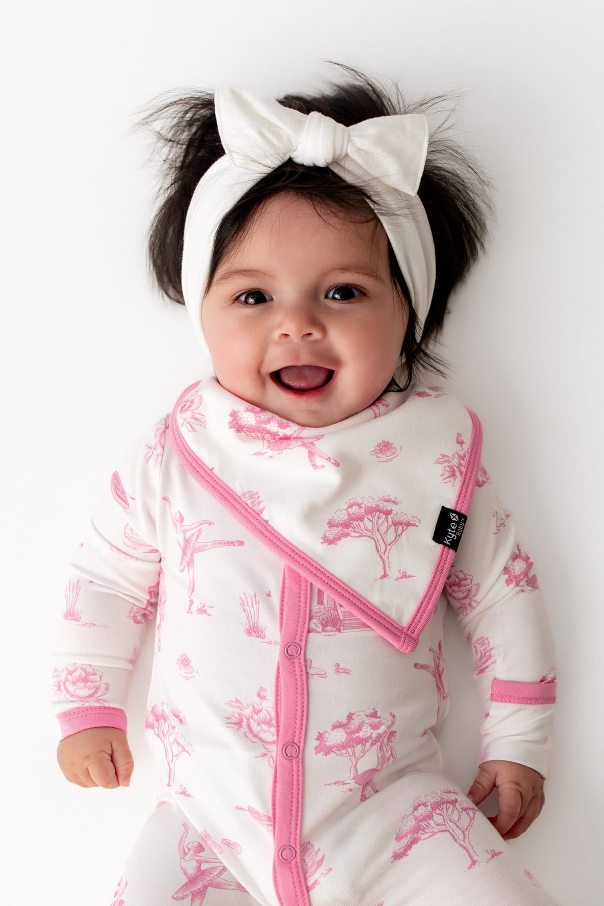 Smiling baby wearing the Bib in Toile Ballet with matching snap romper with a white bow