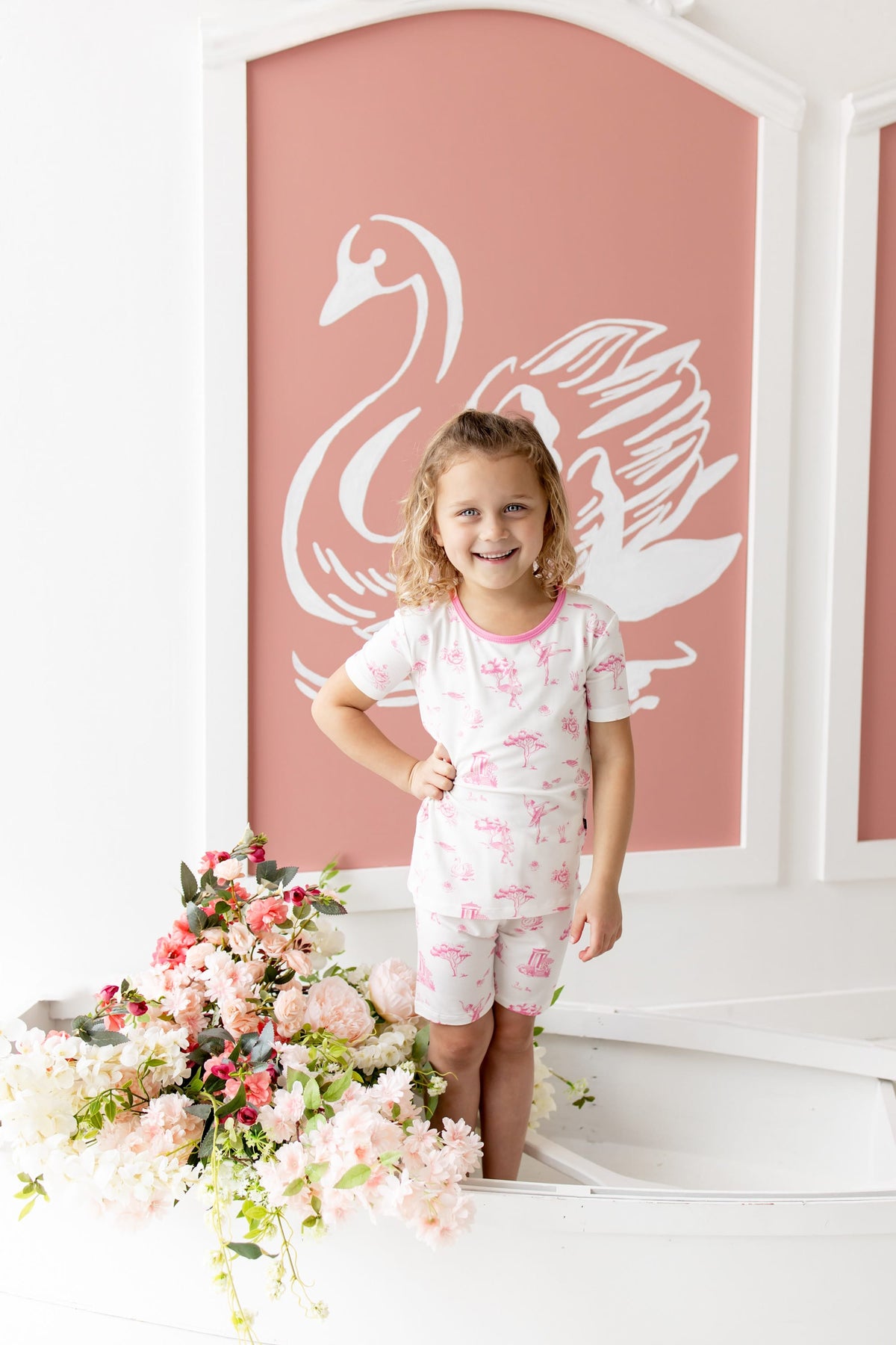 Young girl wearing the Short Sleeve Pajamas in Toile Ballet posing standing inside a white rowboat beside flowers in front of a white swan on a pink background painting