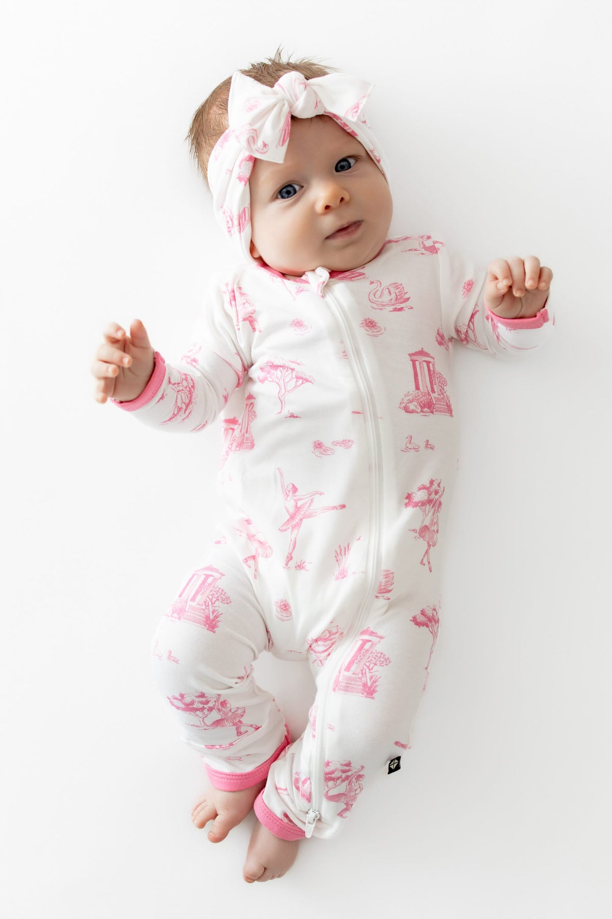 Infant laying on a white surface wearing the Zippered Romper in Toile Ballet and matching bow